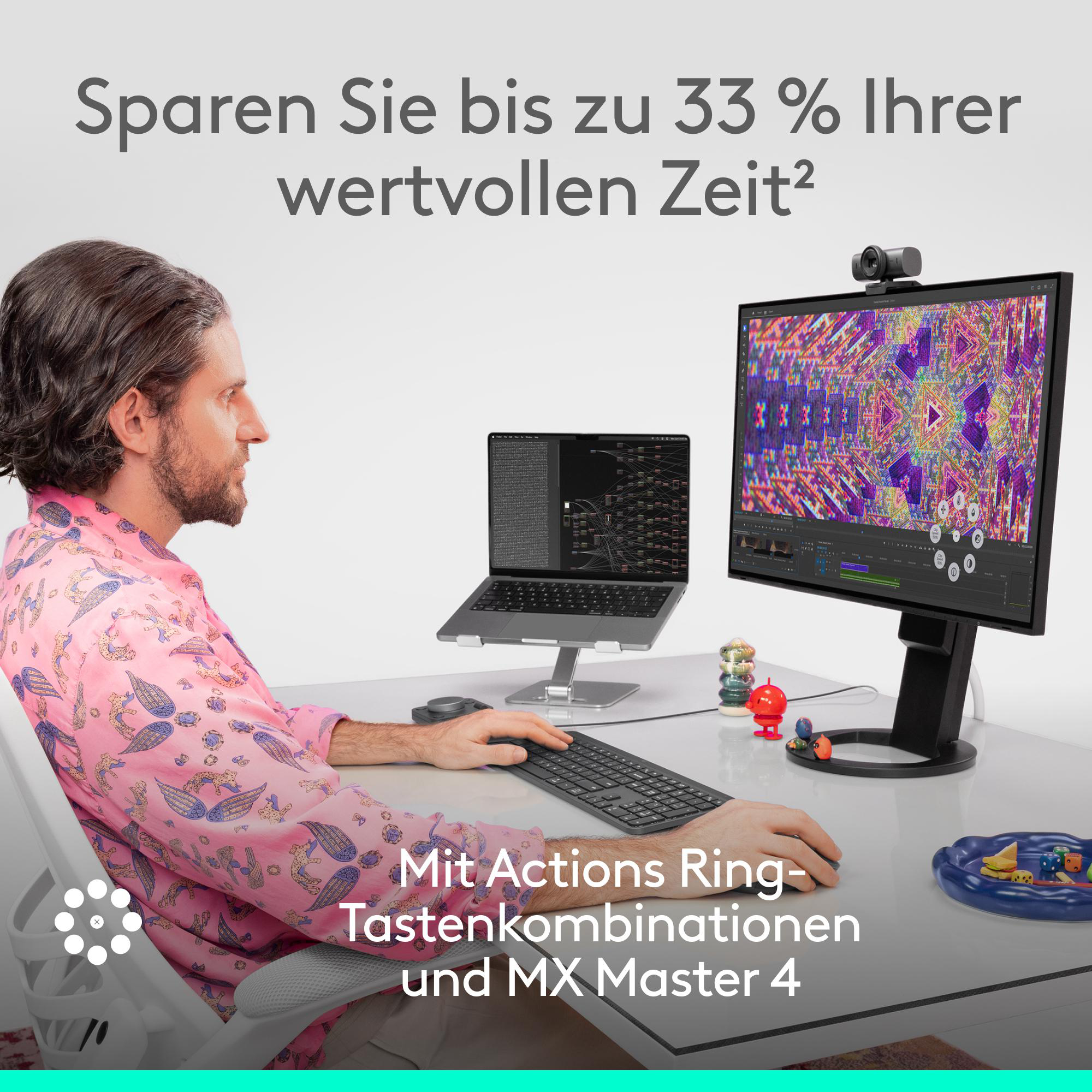 Mann am Schreibtisch mit Laptop, Monitor und Tastatur. Text auf dem Bildschirm und auf Deutsch. Farbige Spielzeuge und Webcam sichtbar.