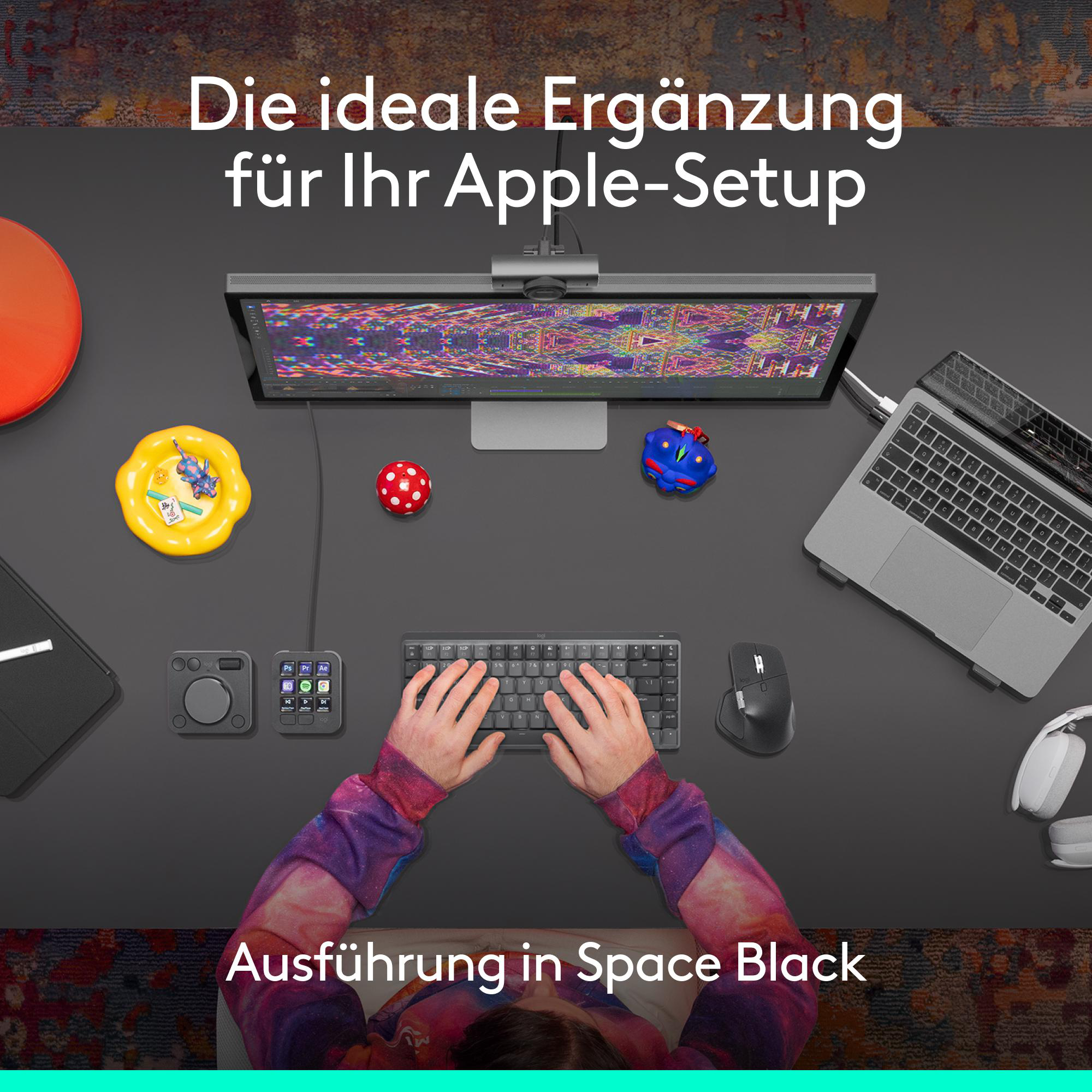 Draufsicht auf einen Arbeitsbereich mit Monitor, Tastatur, Laptop und Zubehör. Hände auf der Tastatur, Text sagt 'Space Black'.