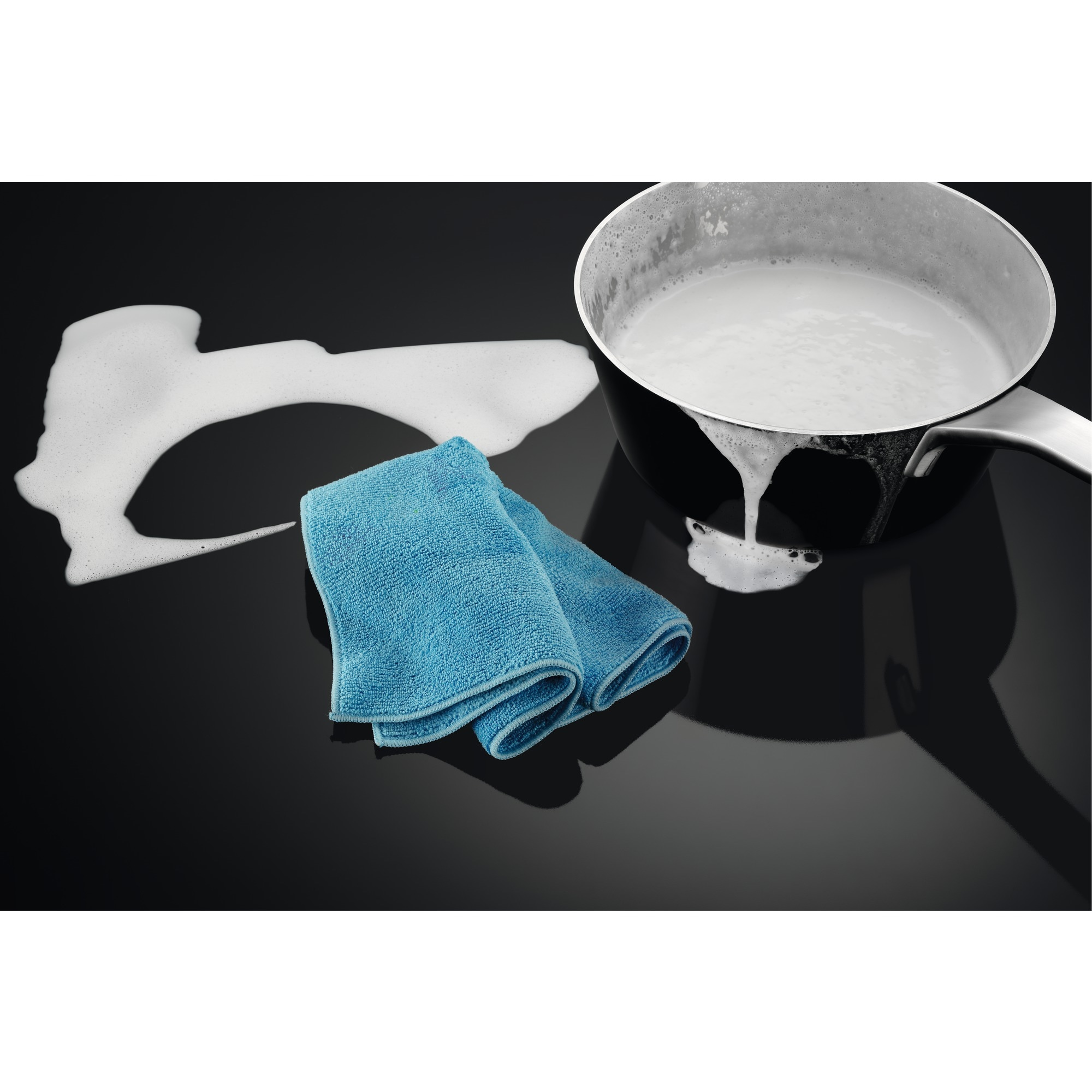 Eine schwarze Kochfeld mit einem Topf mit überlaufender Milch, einem blauen Tuch und verschäumter Milch.