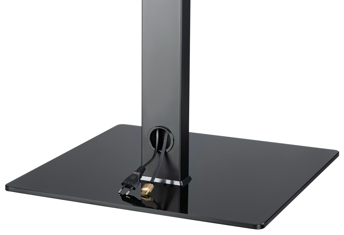 Czarny stojak pod telewizor z kwadratową podstawą i pionowym wspornikiem z otworem. Widoczne kable, podłączone do stojaka.