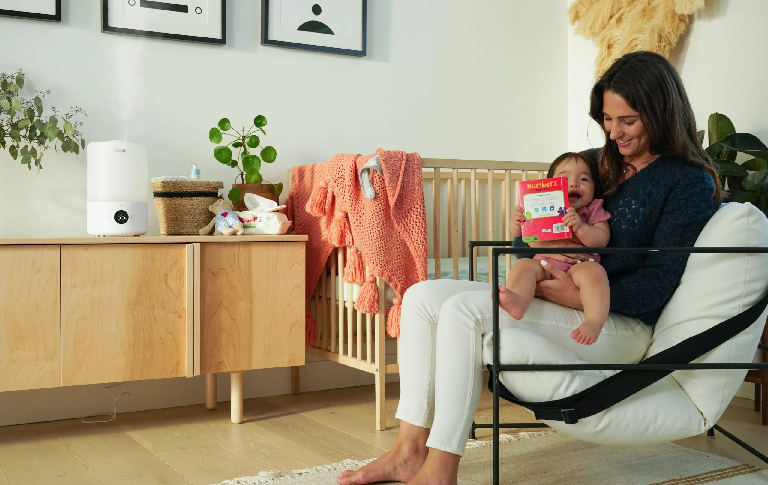 Kobieta trzyma dziecko czytające książkę. W pokoju jest kołyska, nawilżacz i dekoracje.