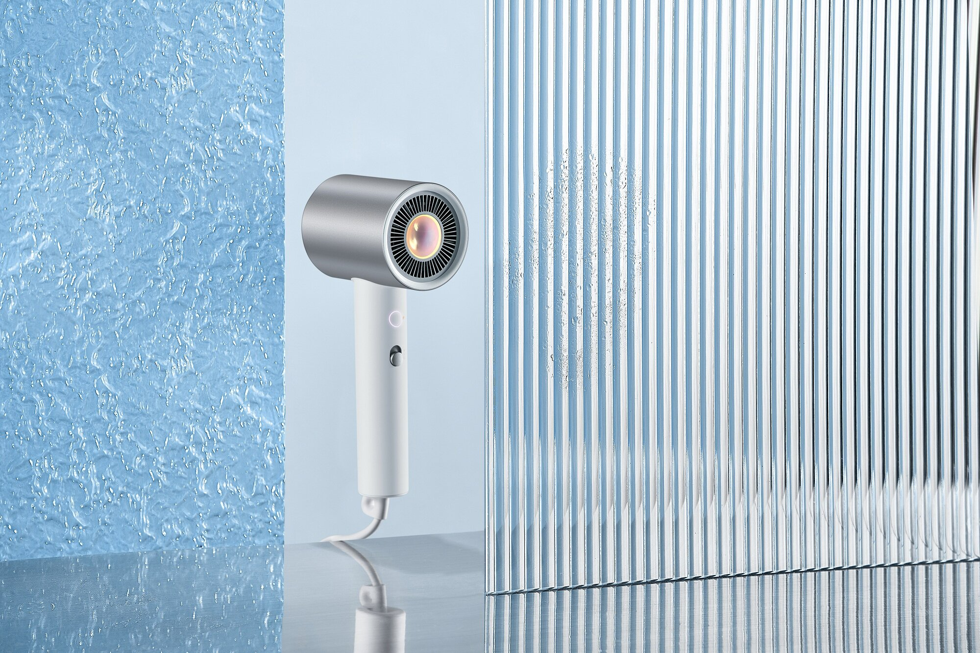 Biała i srebrna suszarka do włosów stoi na odblaskowej powierzchni. Jest na tle niebieskiej tekstury i pionowego panelu.