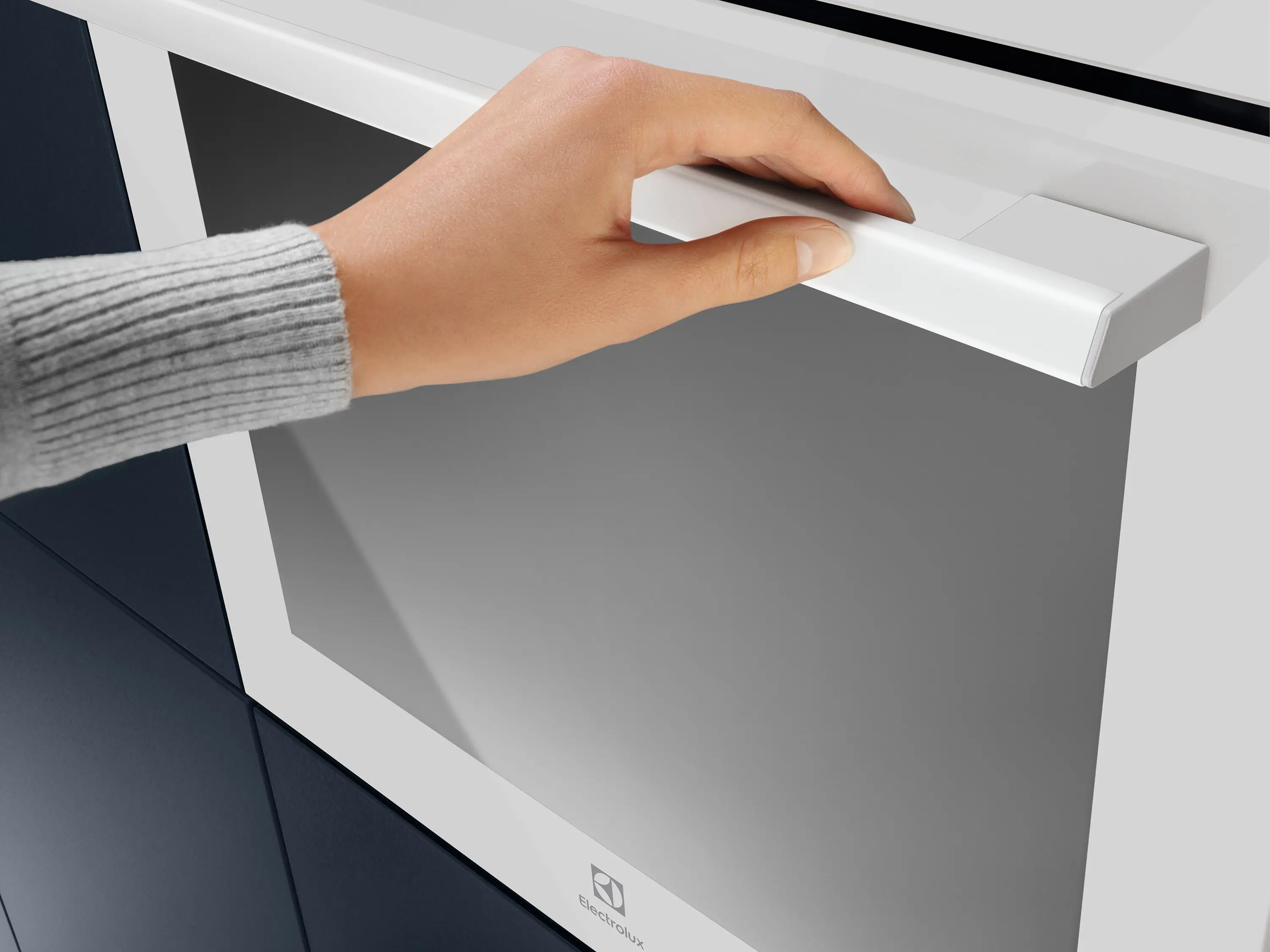 Ręka dotyka uchwytu drzwi piekarnika. Uchwyt jest biały, a drzwi szare. Widoczny jest szary sweter.