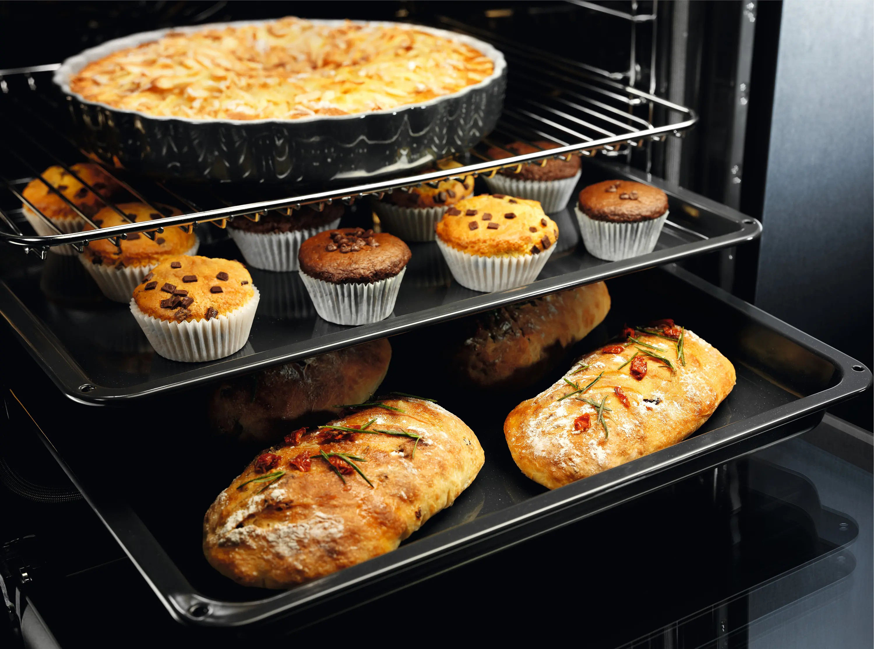 Piekarnik z ciastem, babeczkami i chlebem. Ciasto w okrągłym naczyniu, babeczki w papilotach, chleb na tacy.