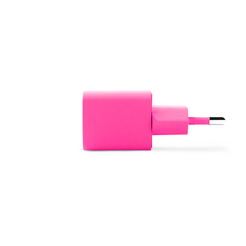 Beyaz bir yüzeyde pembe bir şarj adaptörü duruyor. Kare bir tabanı ve siyah bir konektörlü daha küçük dikdörtgen bir fişi var.