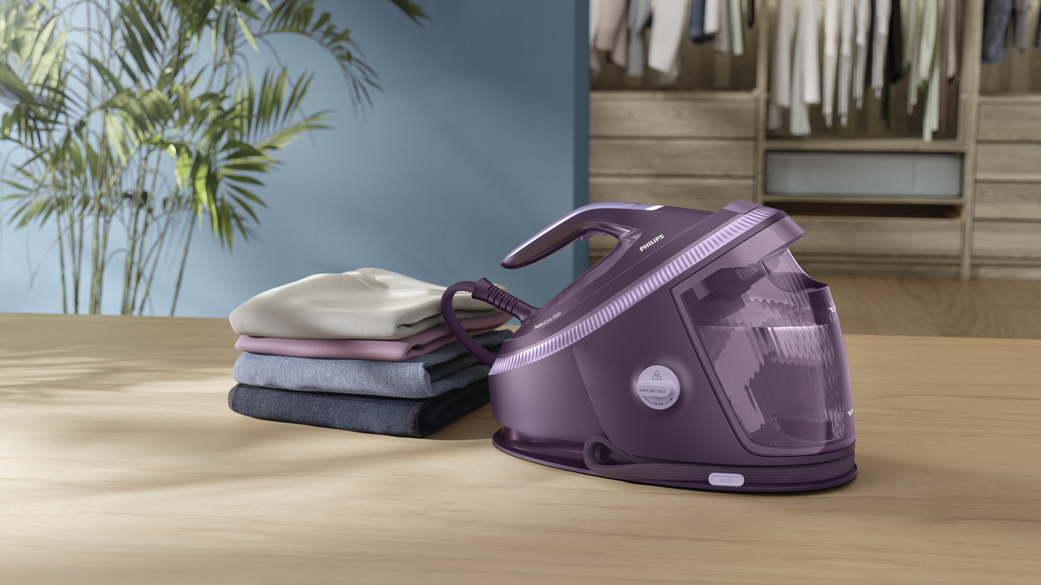 Fioletowe żelazko obok stosu ubrań na drewnianym stole. Po lewej roślina, w tle garderoba.