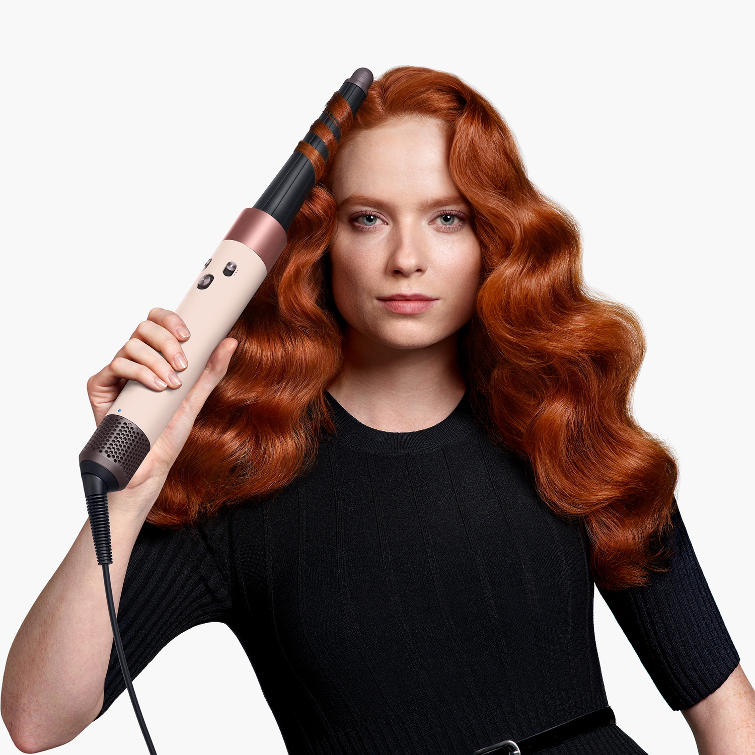 Woman with red hair holding a curling iron. She wears a black dress. White background.