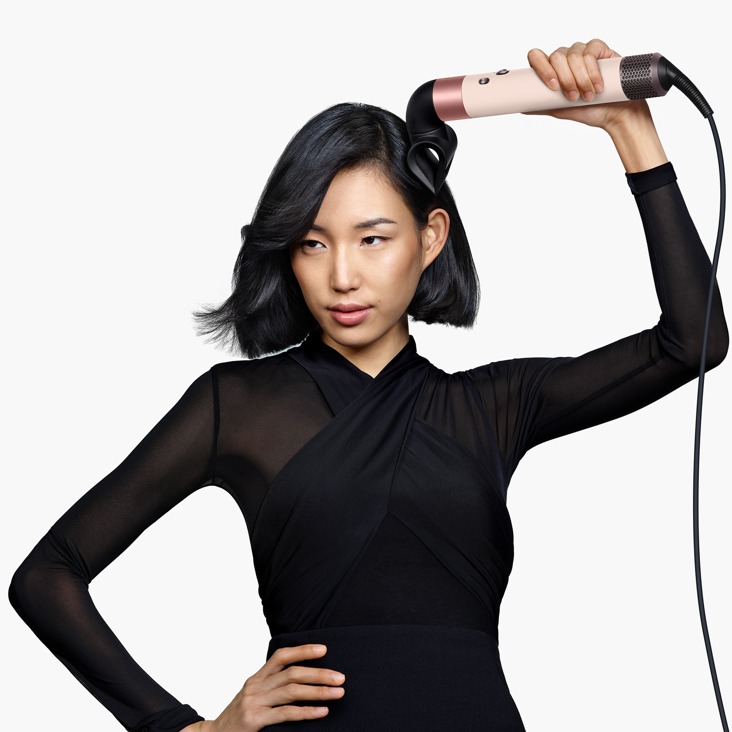 Woman styling hair with a hair styler. She wears a black top and skirt. The background is white.