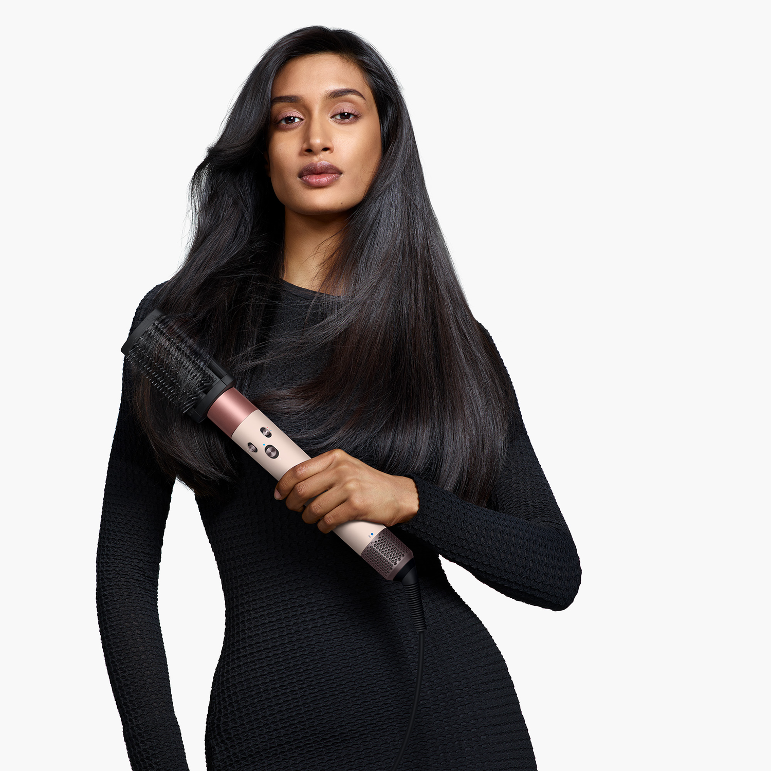 A woman with long black hair holds a hair styling tool. She wears a black dress, and the background is white.
