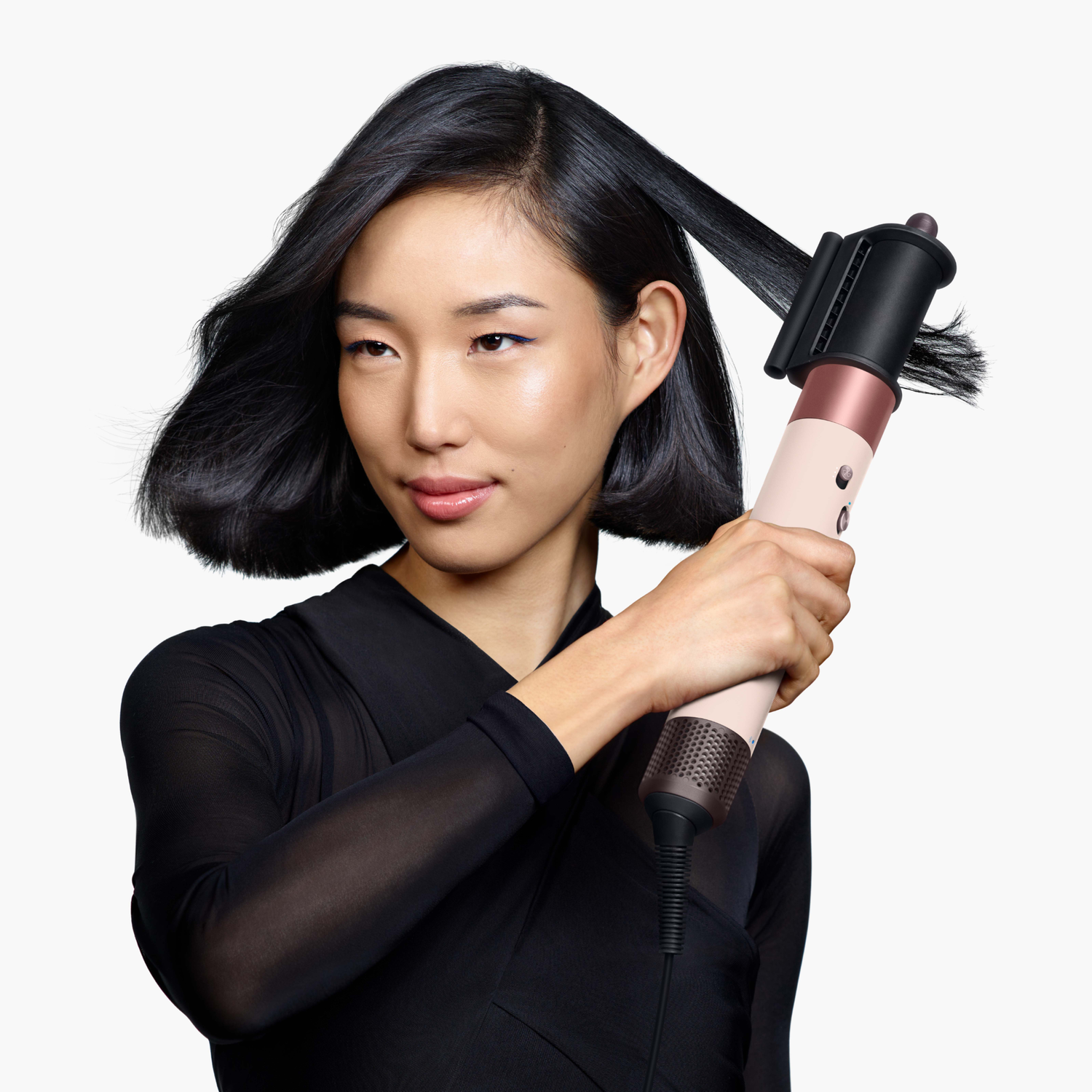 A woman with black hair uses a hair styler. She wears a black top. The styler is pink, black, and copper-colored, with hair in it.