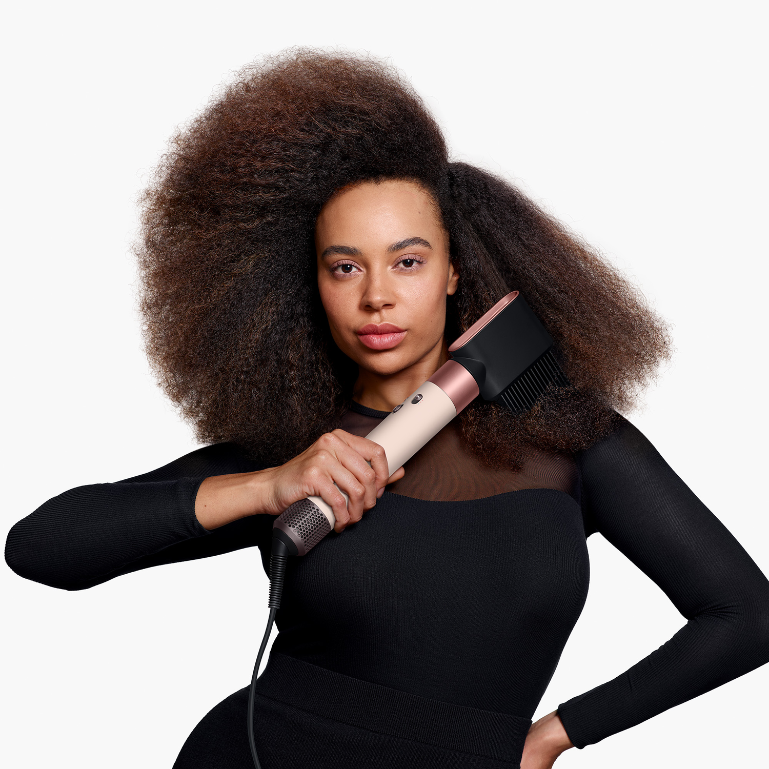 A woman with voluminous hair holds a hair dryer. She wears a black dress. The background is white.