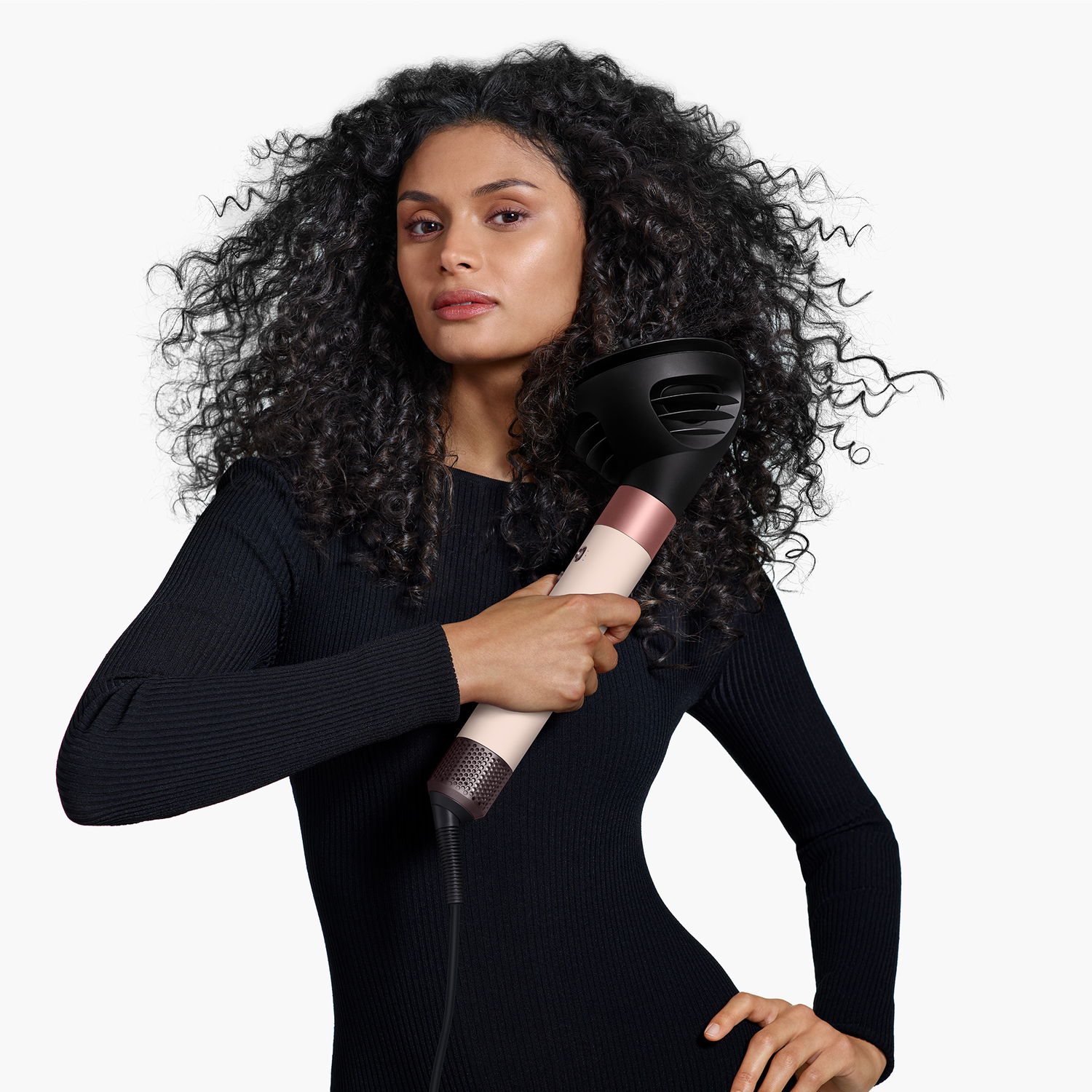 A woman with curly hair holds a hairdryer. She wears a black dress. The background is white.