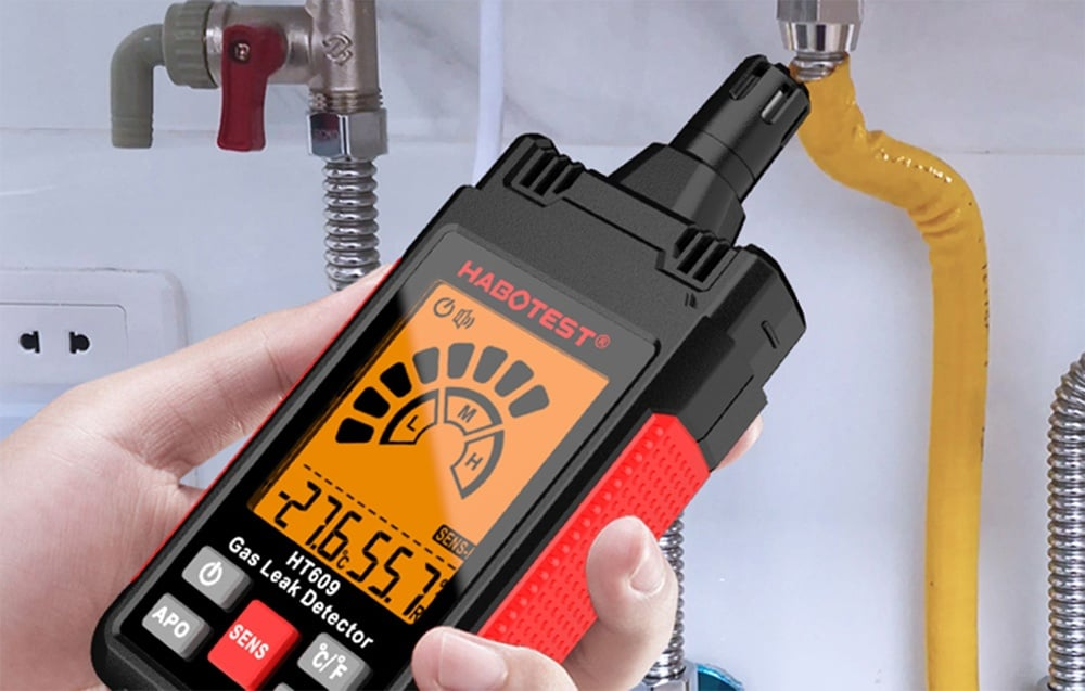 Egy kéz gázszivárgás-érzékelőt tart, amely leolvasásokat és egy mérőműszert mutat. A háttérben csövek, egy szelep és egy elektromos aljzat látható.