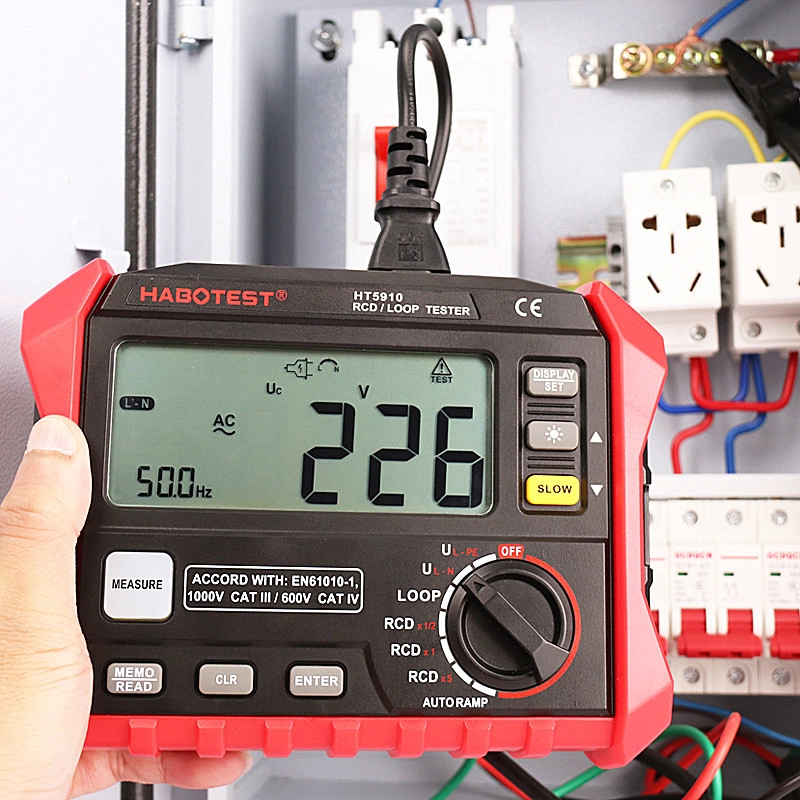 Egy kéz egy piros-fekete villamosmérőt tart, ami 226 V-ot mutat. A háttérben más elektromos alkatrészek és vezetékek láthatók.