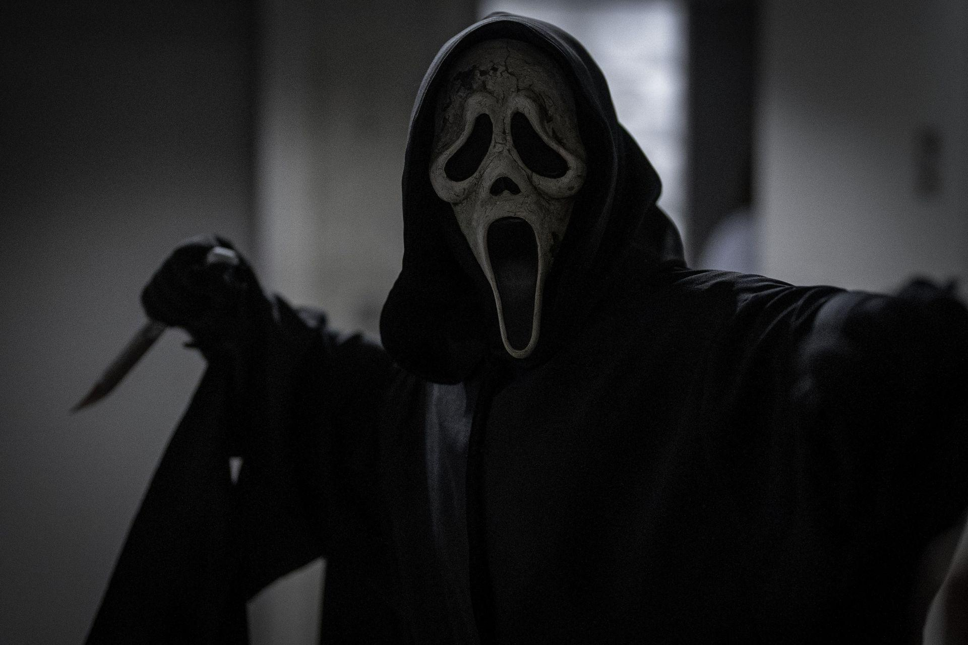 Eine Person mit Ghostface-Maske und Kapuzenrobe hält ein Messer. Die Maske ist weiß mit offenem Mund, in einem dunklen Raum.