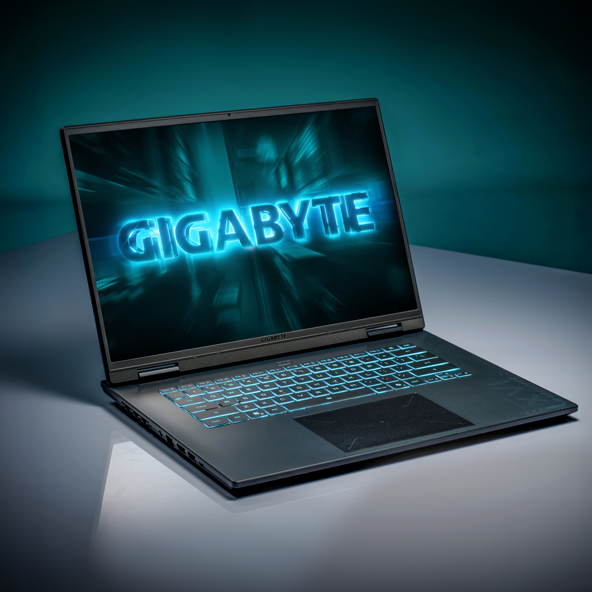 Ein Laptop zeigt das Wort GIGABYTE in Blau, mit einer hintergrundbeleuchteten Tastatur. Der Hintergrund ist türkis und die Oberfläche reflektierend.