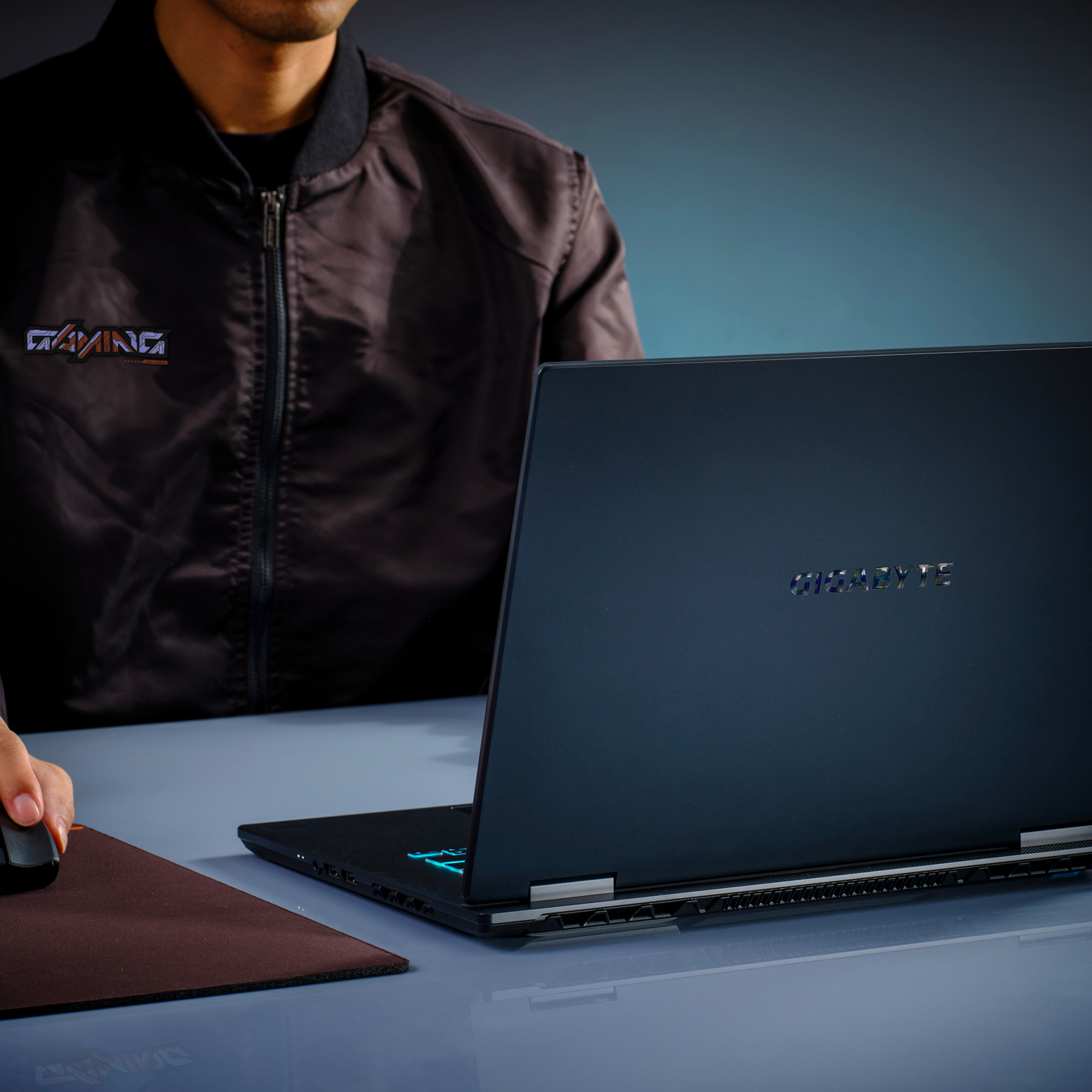 Eine Person benutzt einen schwarzen Laptop mit GIGABYTE-Logo. Sie trägt eine schwarze Jacke und benutzt eine Maus auf einem braunen Pad.