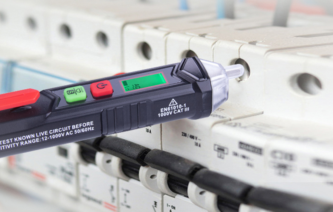 Egy feszültségmérő ellenőrzi az elektromos panelt. A teszter fekete, zöld és piros gombokkal és digitális kijelzővel rendelkezik. A panel fehér.
