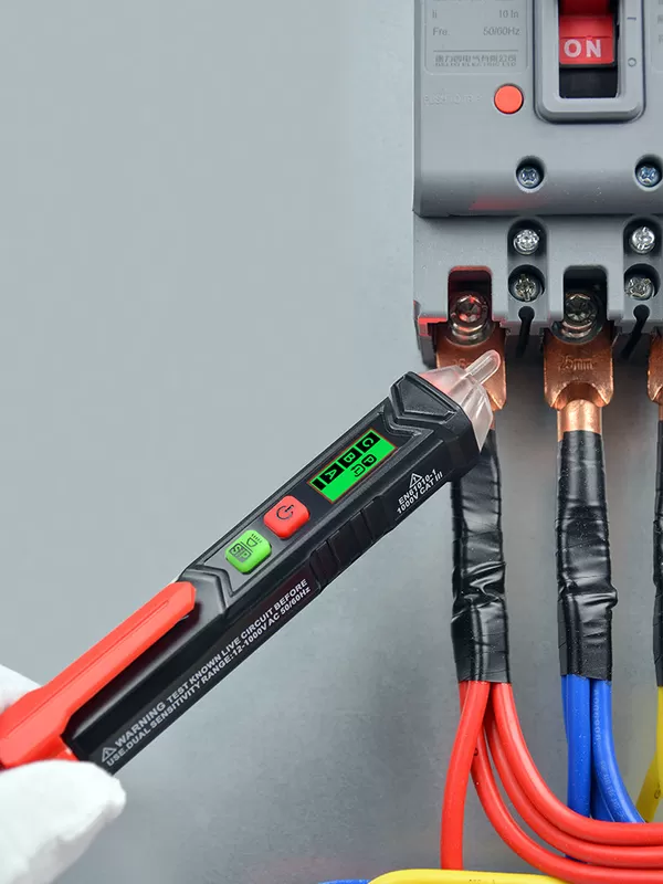 Egy fekete-piros feszültségmérőt tartanak elektromos vezetékek és egy megszakító közelében. A képernyő zöld fényt mutat. A megszakító 'ON' felirattal van ellátva.