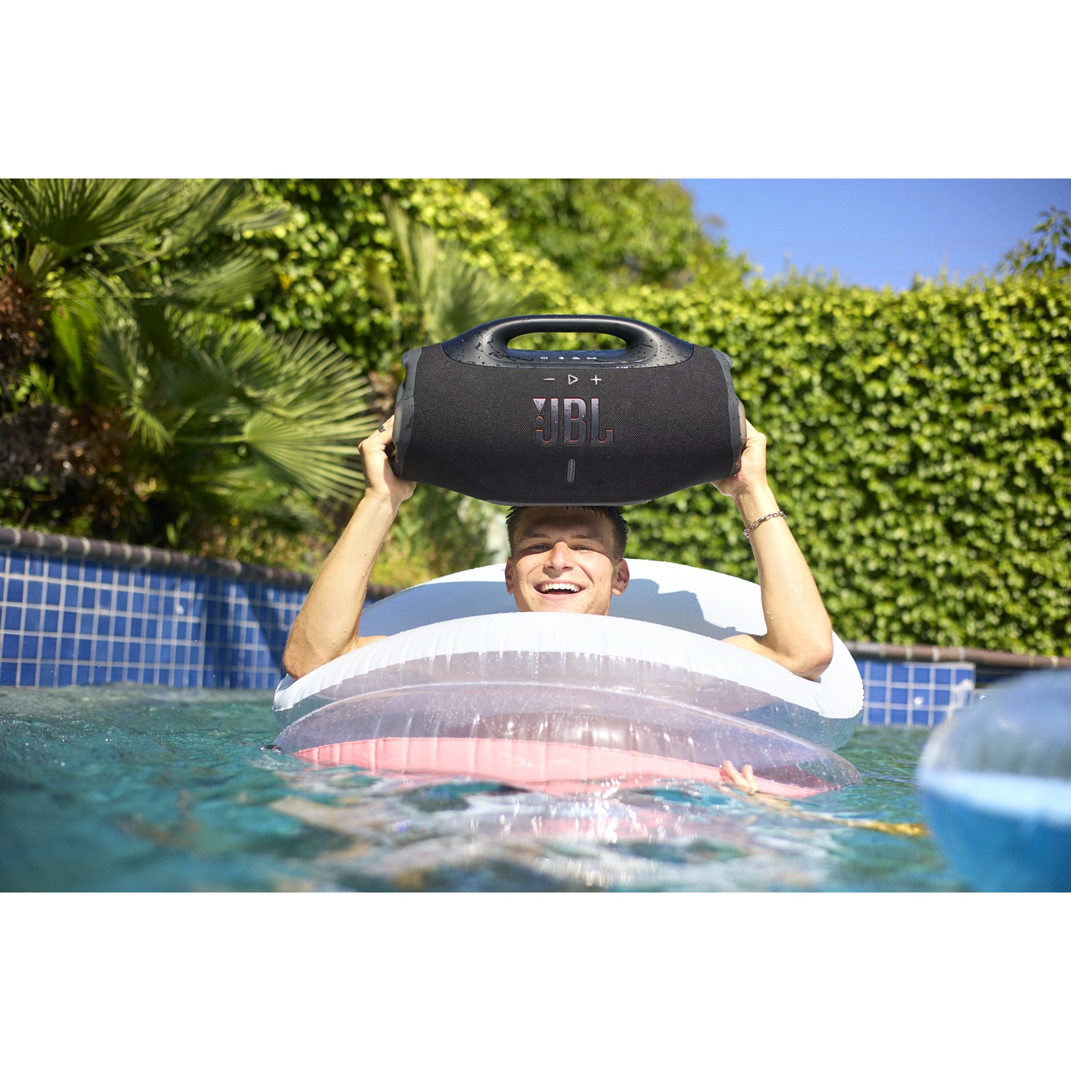 A man in a pool holding a JBL speaker above his head. He is in a white and pink inflatable ring, smiling.