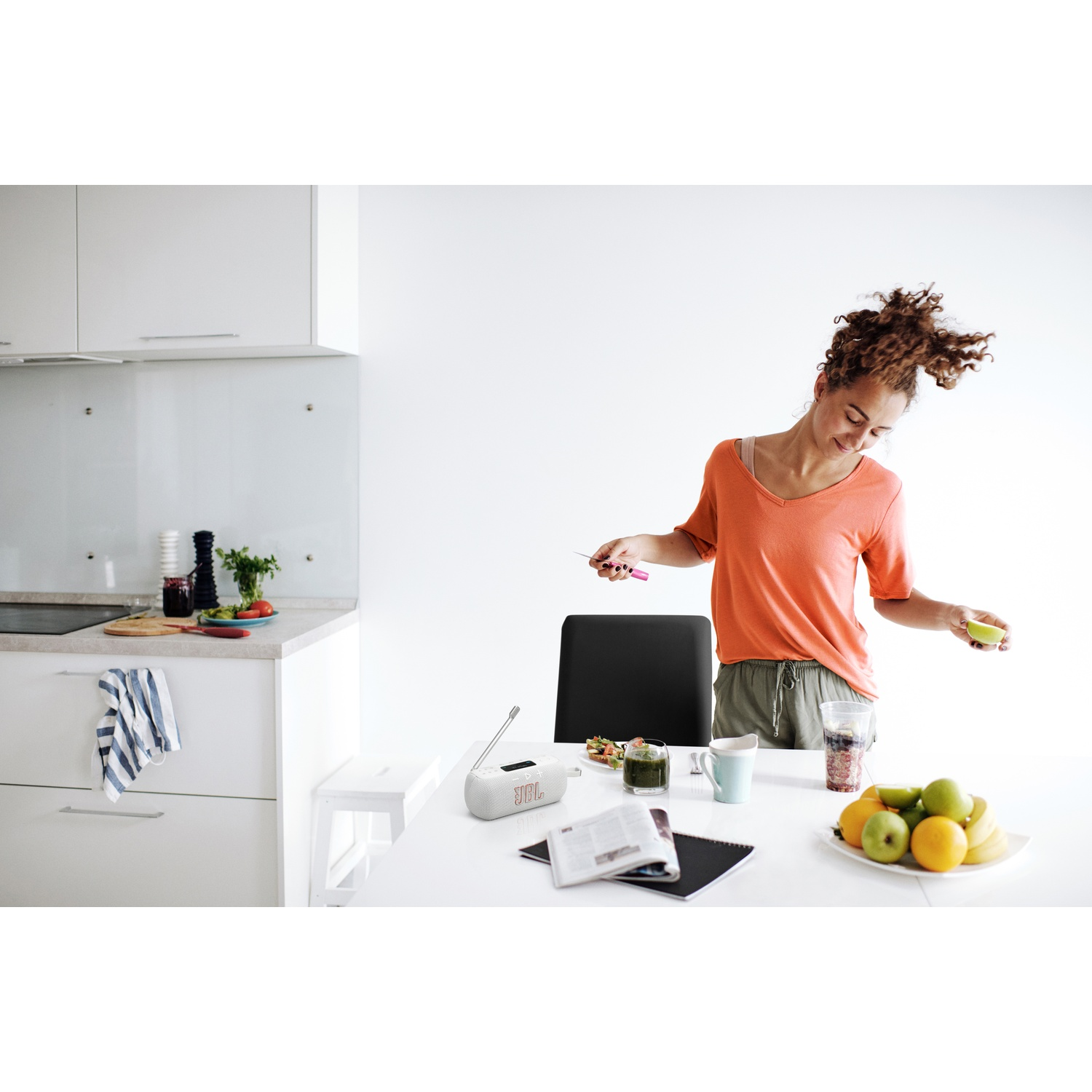 Woman in orange shirt in a kitchen. She holds a lime and a small tool. A radio, fruit, and drinks are on the table.