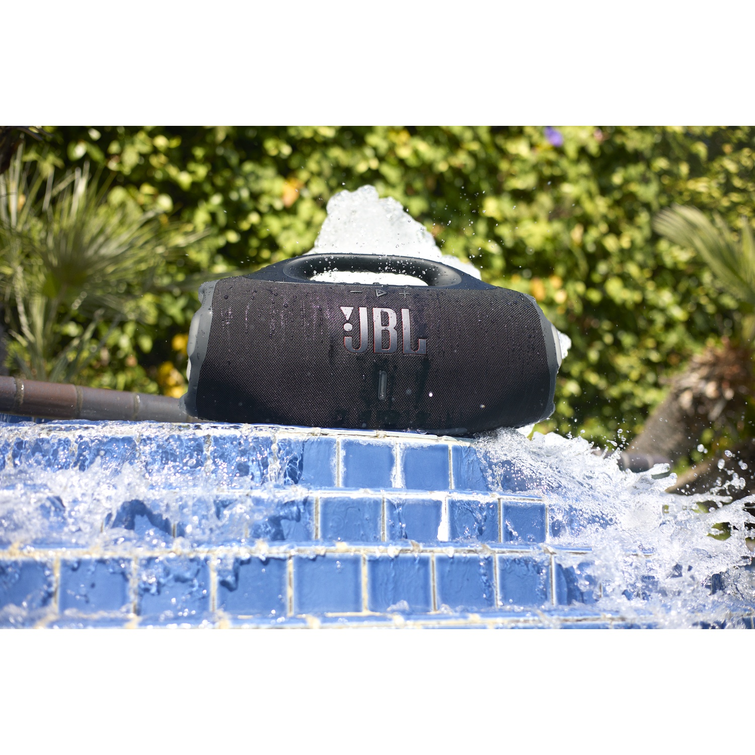 A black JBL speaker sits on blue tiles as water splashes around it. Green foliage and a fountain are in the background.