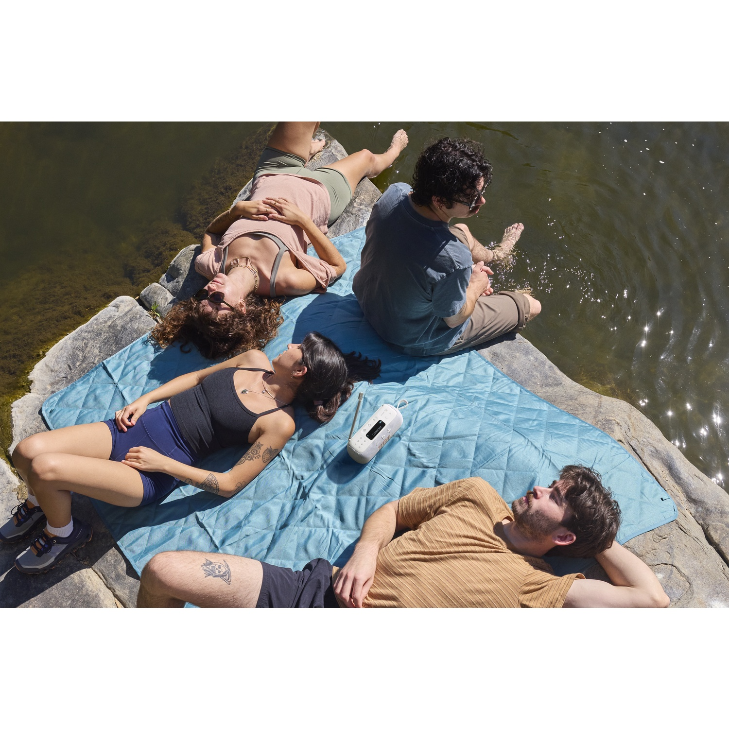 Four people relaxing on rocks and a blue blanket by water. One is in the water. A speaker sits on the blanket.