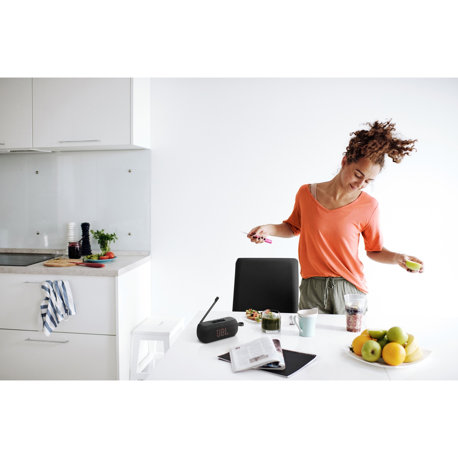 Vrouw in oranje top, eten aan het bereiden. Keuken met tafel, JBL-luidspreker, fruit en groenten. Heldere, witte achtergrond.