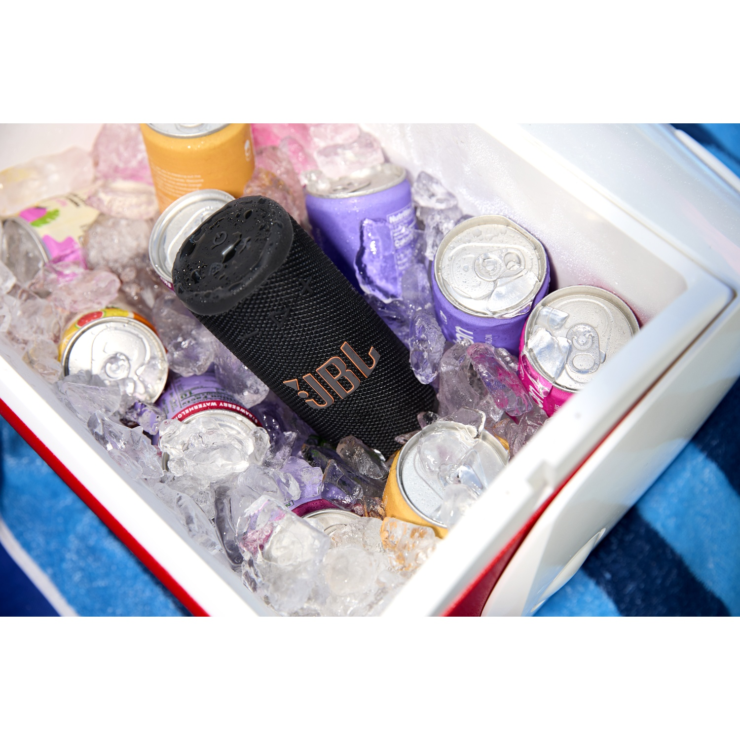A cooler filled with ice and cans of soda and drinks, a black JBL speaker, and a blue striped towel in the background.