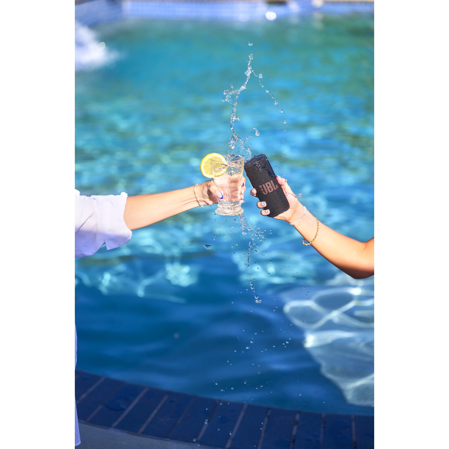Two hands hold a glass of water with lemon and a JBL speaker, water splashing in front of a swimming pool.