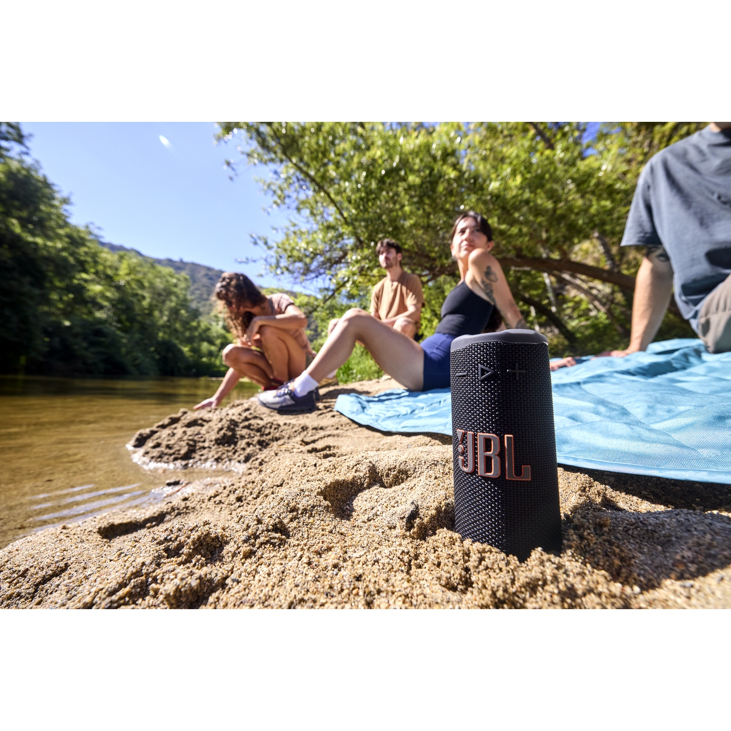 A black JBL speaker sits on sand. People are relaxing by a river on a sunny day. One person touches the water.