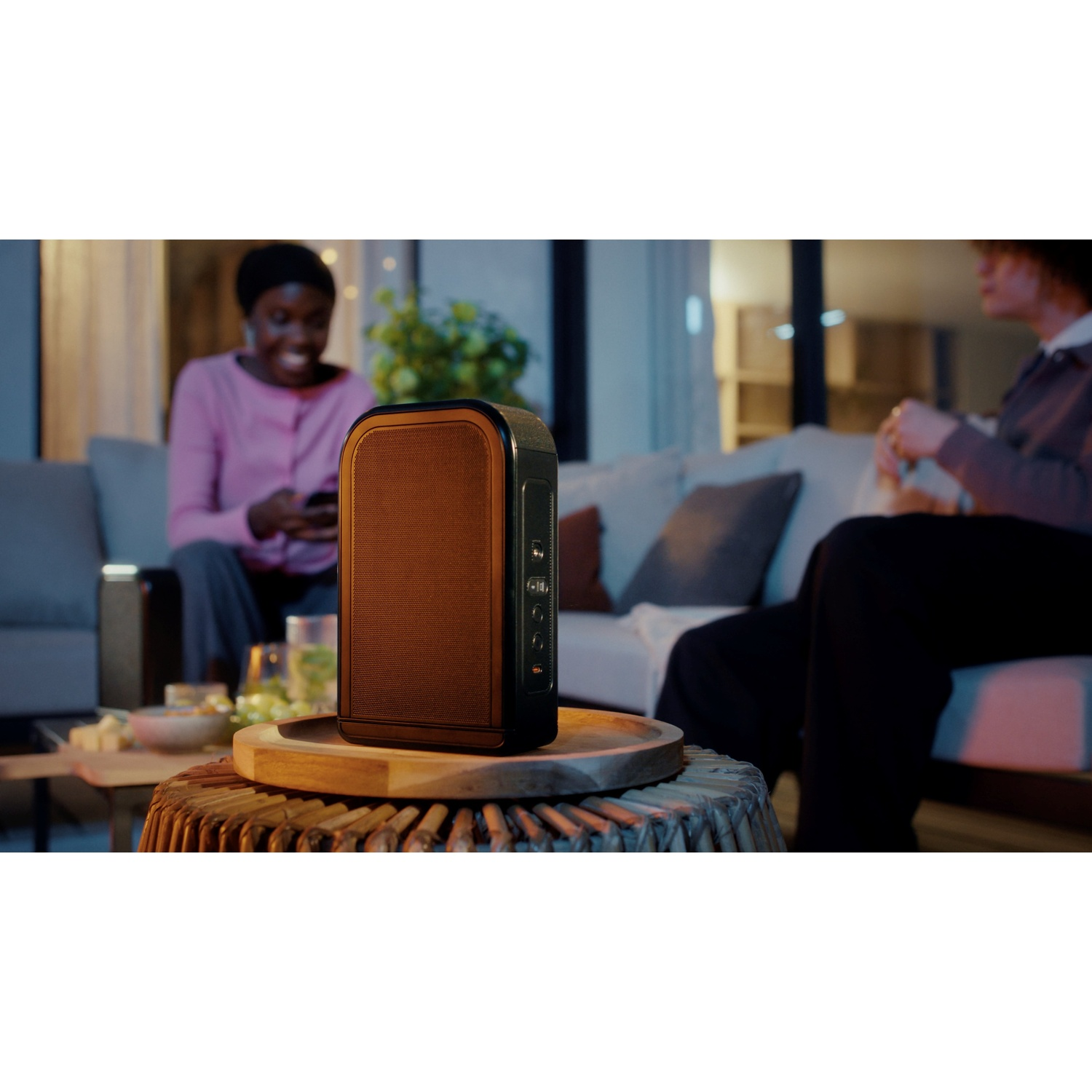 Un haut-parleur est posé sur une table. Des personnes sont en arrière-plan, l'une utilise un téléphone, l'autre est assise. Cadre extérieur.