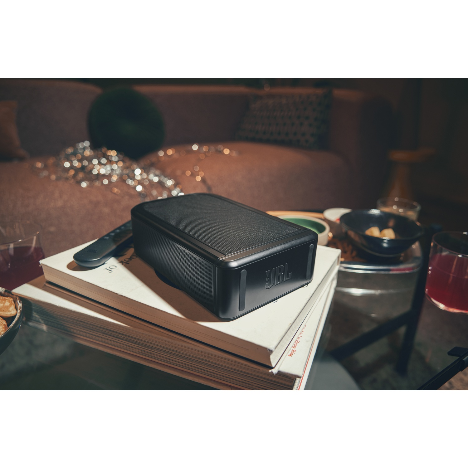 Un haut-parleur JBL noir est posé sur des livres avec une télécommande, près de boissons et de collations.