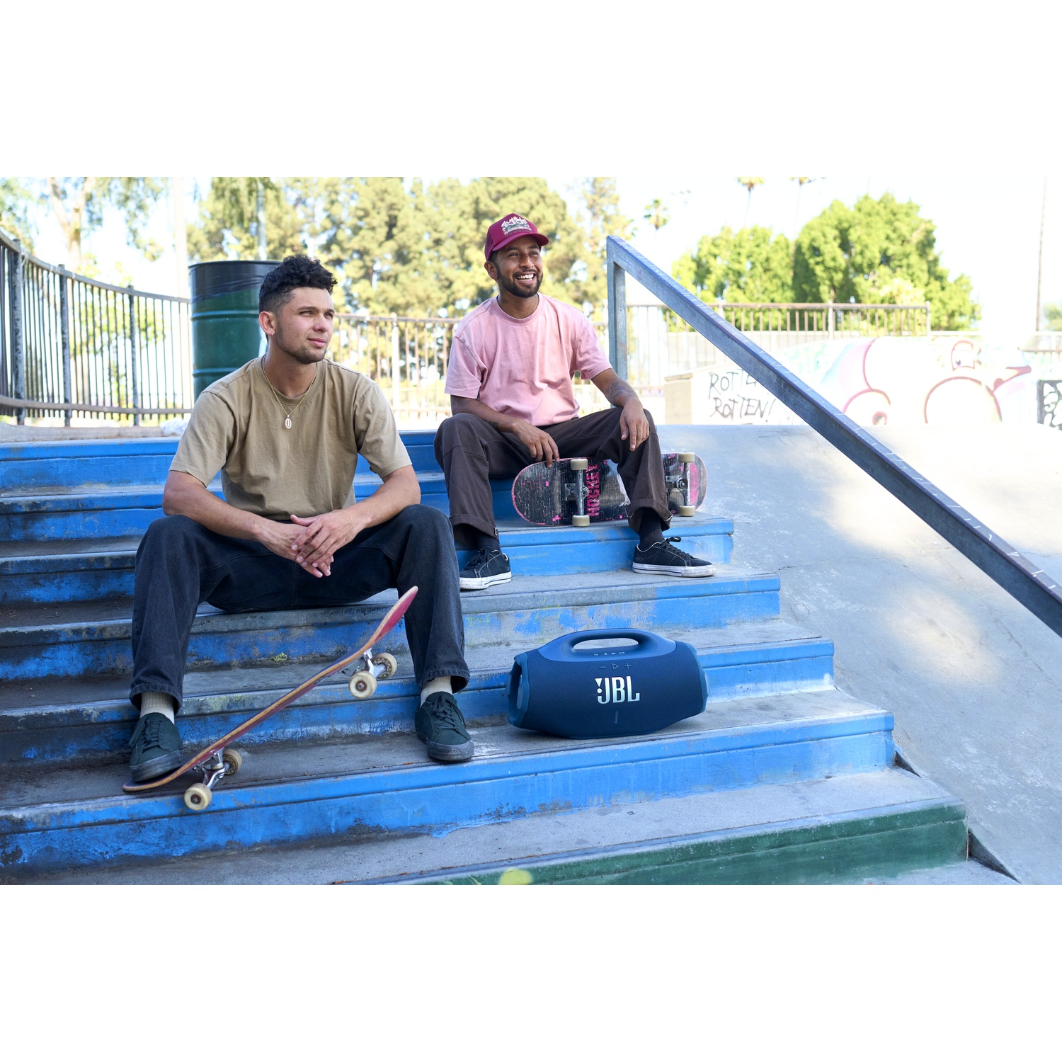 Deux hommes assis sur des escaliers bleus dans un skatepark, l'un avec un skateboard, l'autre avec un skateboard et un haut-parleur JBL.