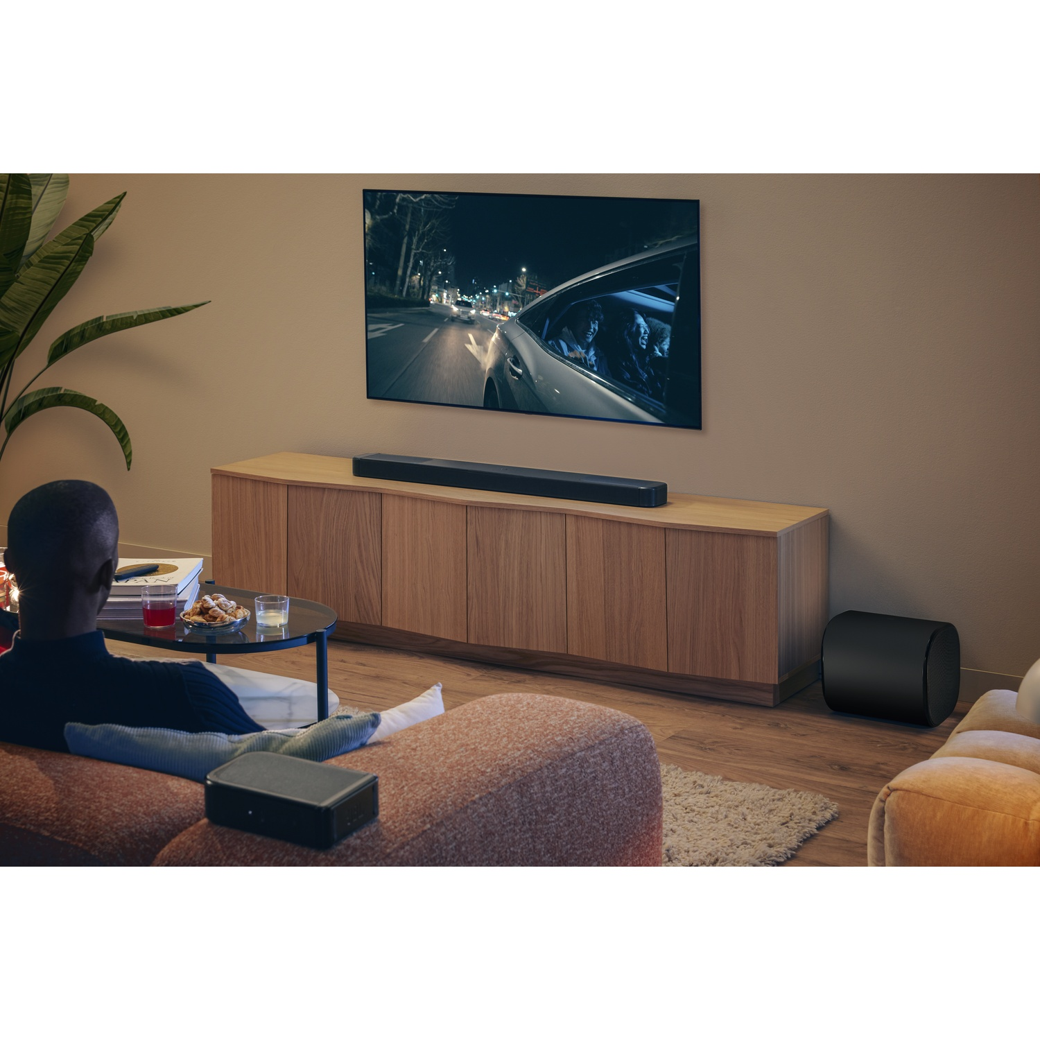 Un homme regarde la télévision. L'écran montre une voiture la nuit. Une barre de son est sous la télé. Un caisson de basses est à droite.