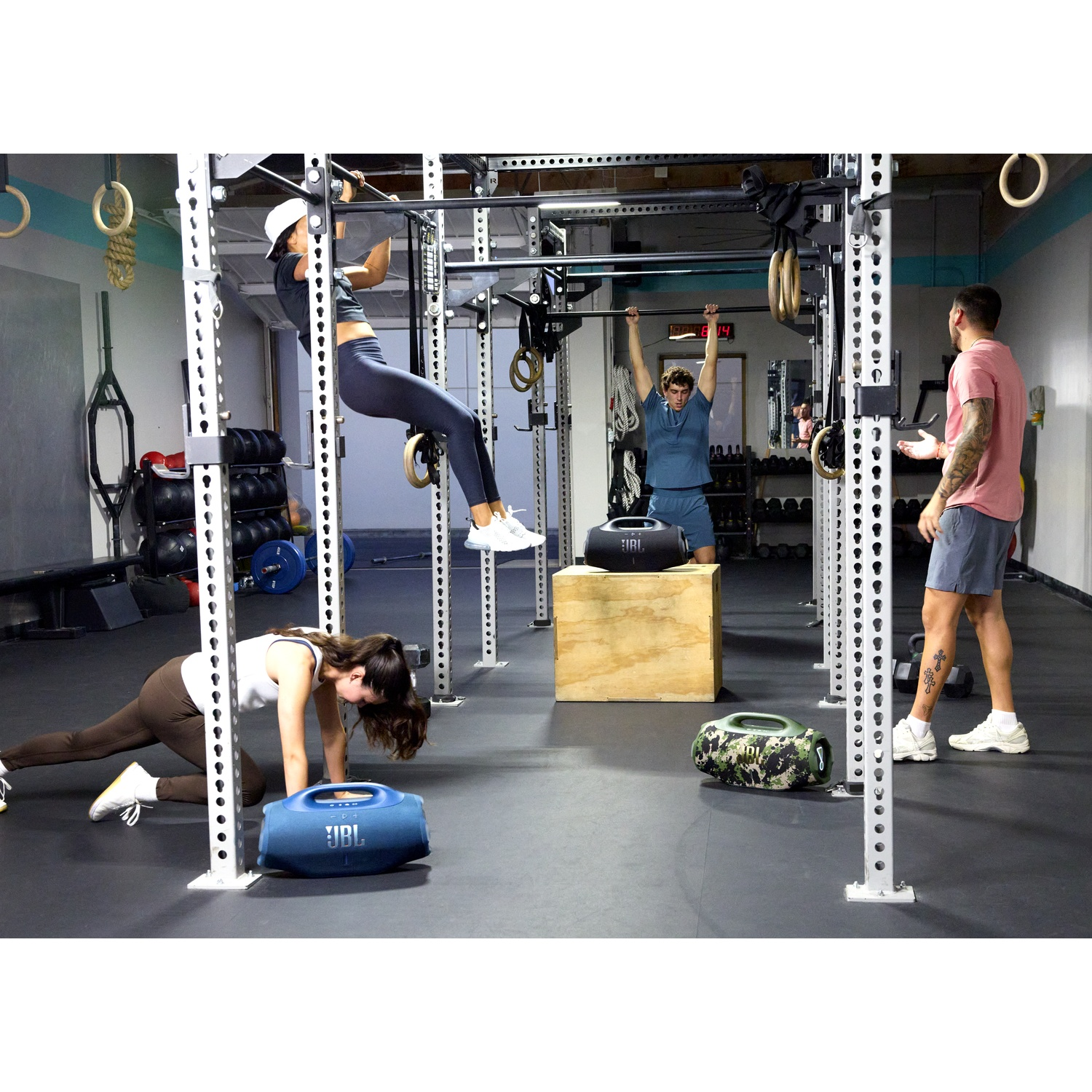 Des personnes s'entraînent dans une salle de sport. Une personne fait des tractions, une autre s'étire et une troisième observe. Des enceintes JBL sont dans la pièce.