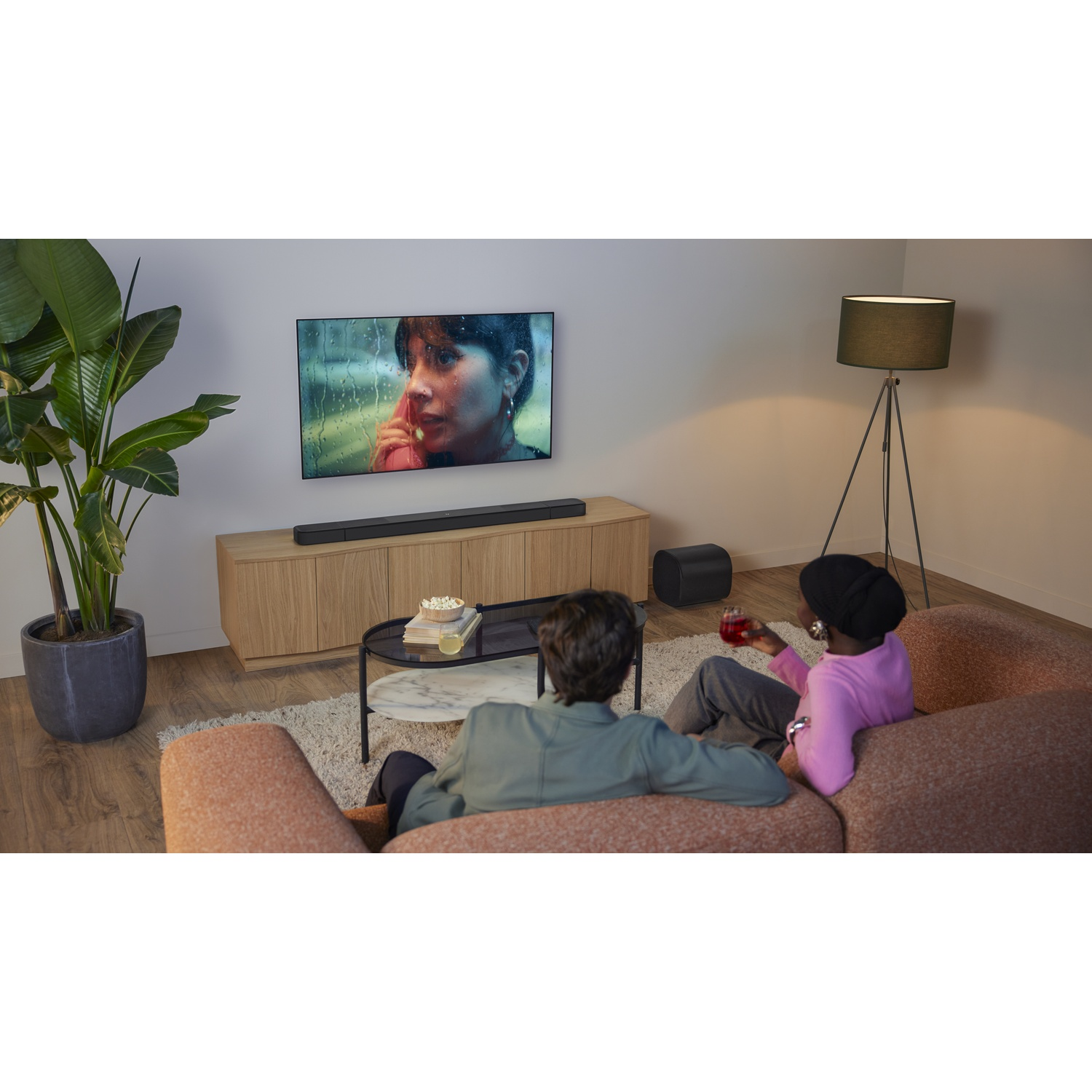 Deux personnes sur un canapé regardent la télévision. Une barre de son est sous la télévision. Une plante et une lampe sont également dans la pièce.