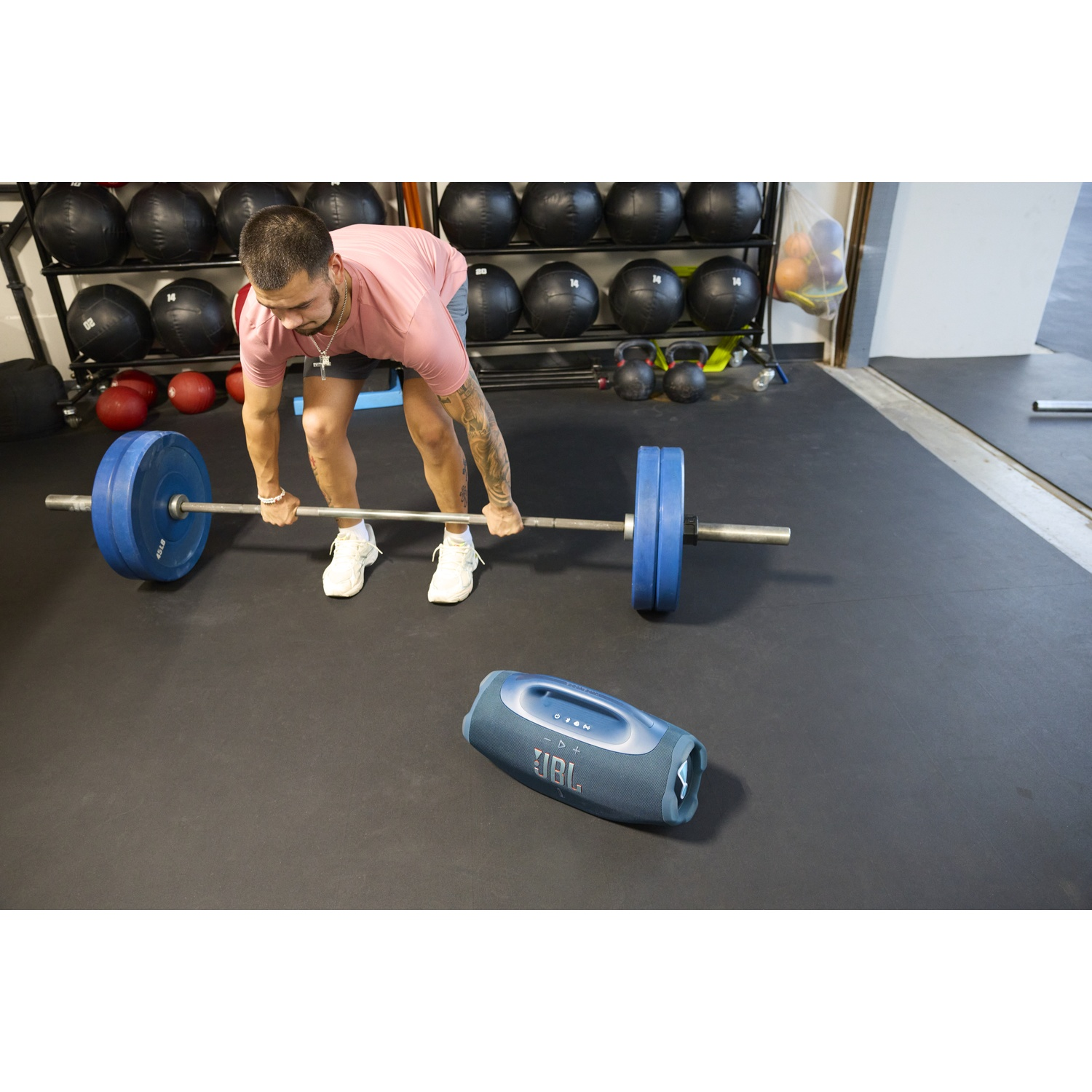 Un homme soulève une barre avec des poids bleus dans une salle de sport. Une enceinte JBL bleue est au sol. L'arrière-plan comprend des équipements de gym.