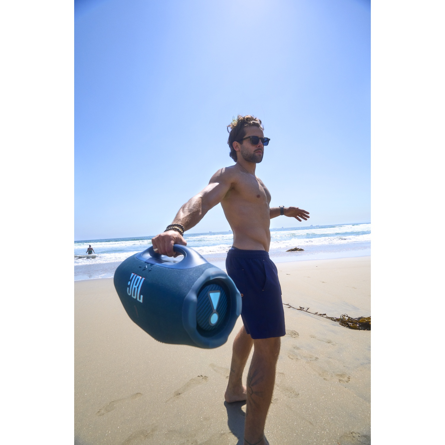 Homme sur la plage tenant une enceinte JBL, portant des lunettes de soleil et un short. Océan et ciel bleu en arrière-plan.