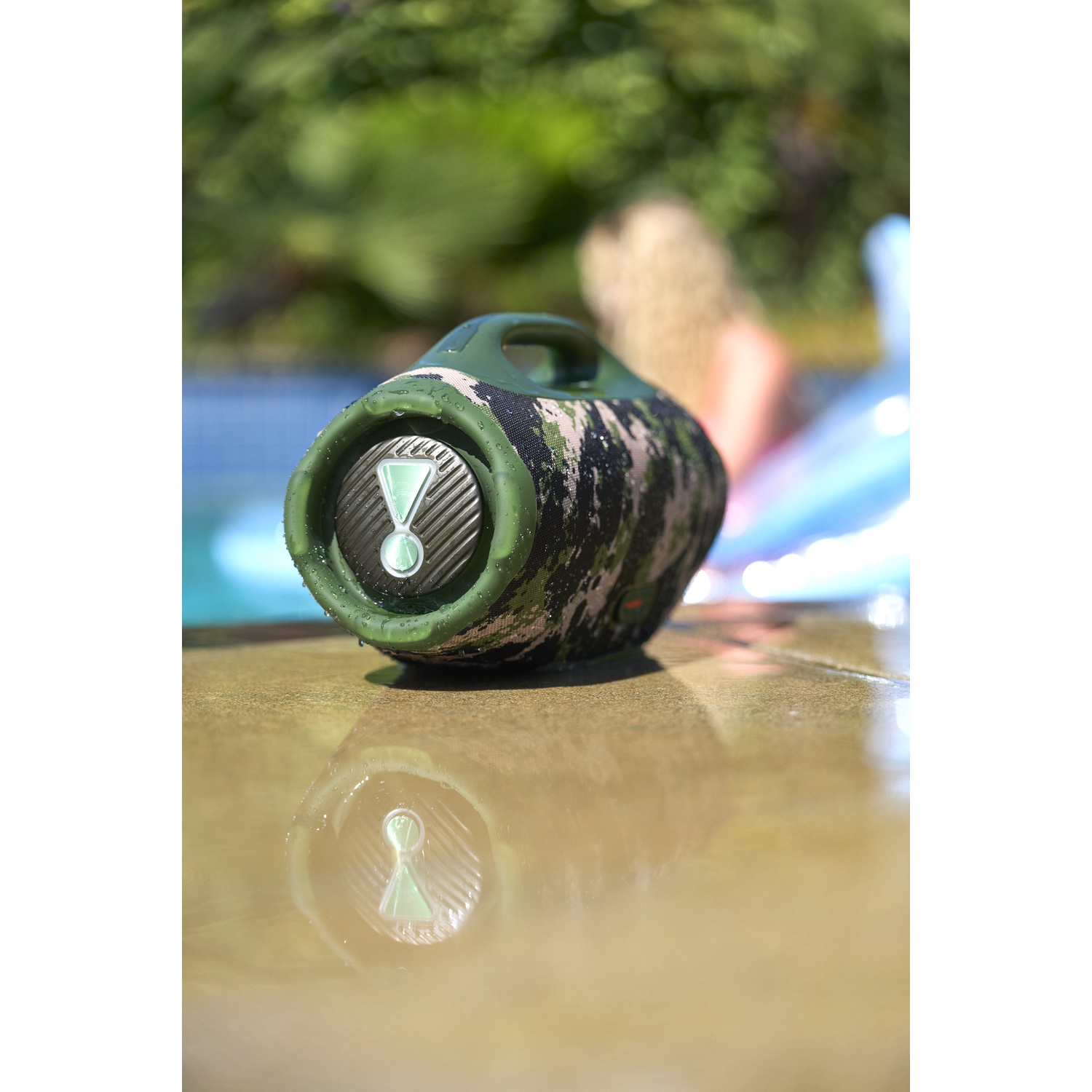 Un haut-parleur camouflage est posé près d'une piscine. Une personne est en arrière-plan. Le haut-parleur se reflète dans l'eau.