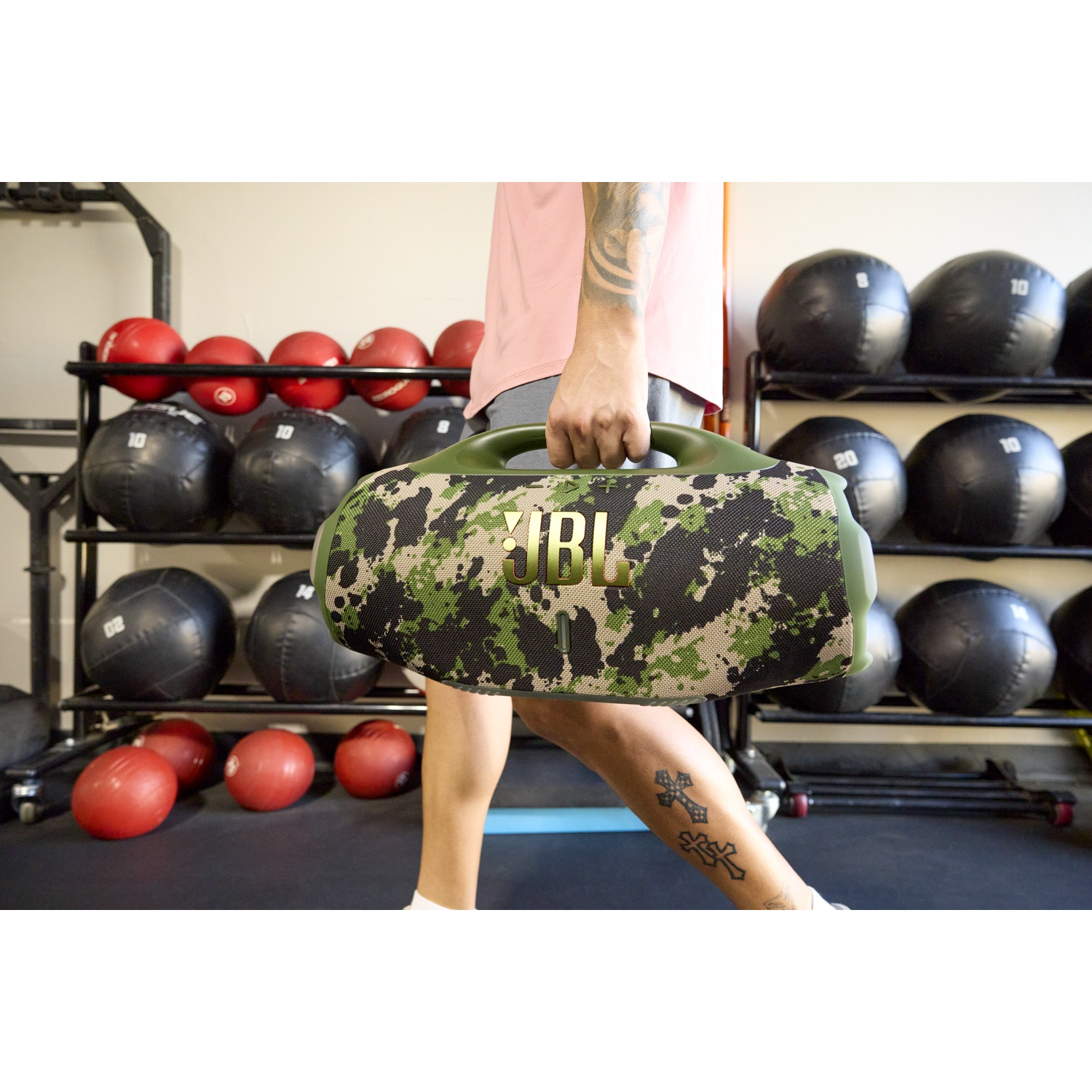 Une personne porte un haut-parleur JBL camouflage. Des équipements de gym et ballons médicaux sont à l'arrière-plan. La personne a un tatouage sur la jambe.