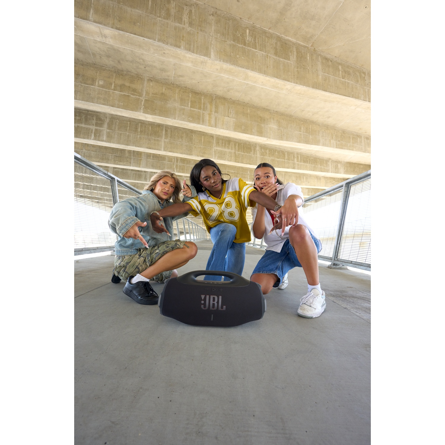 Three people pose with a JBL speaker. The background shows a concrete structure. They are pointing at the camera, dressed in casual wear.