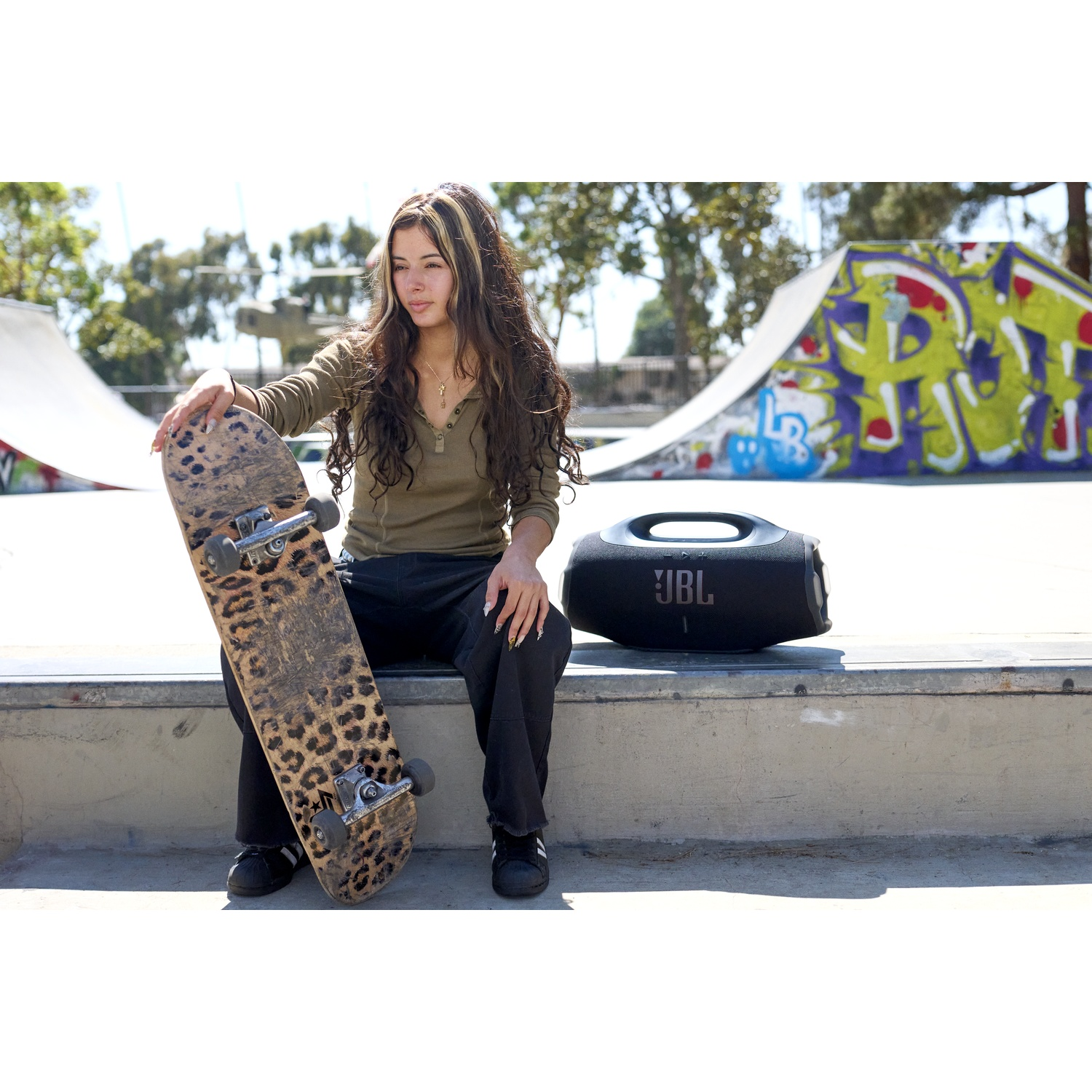 A young woman sits with a leopard print skateboard. A black JBL speaker sits next to her. A skatepark ramp is in the background.