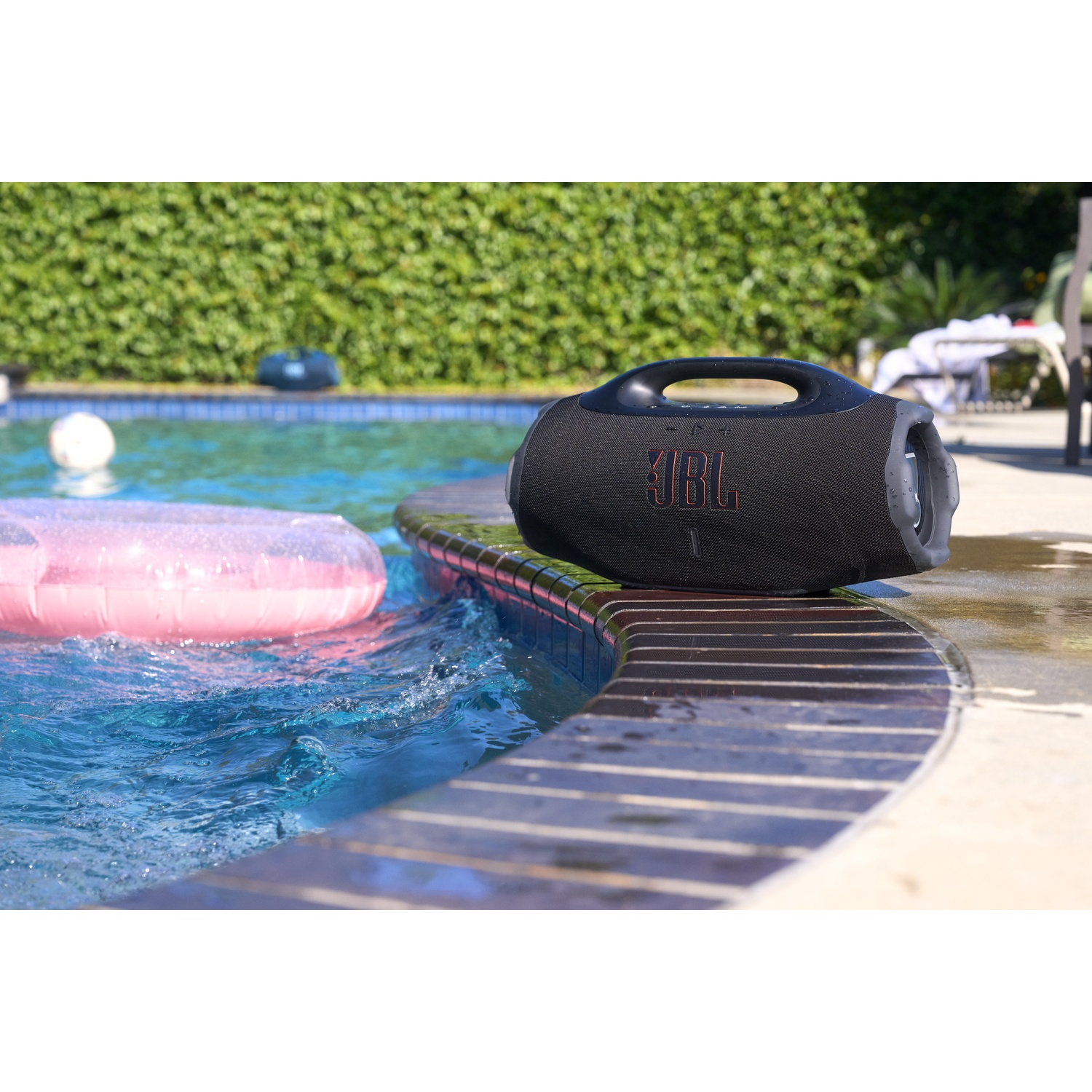 A black JBL speaker rests on a pool's edge. Pink inflatable, blue water, green bushes in background. Sunny day.