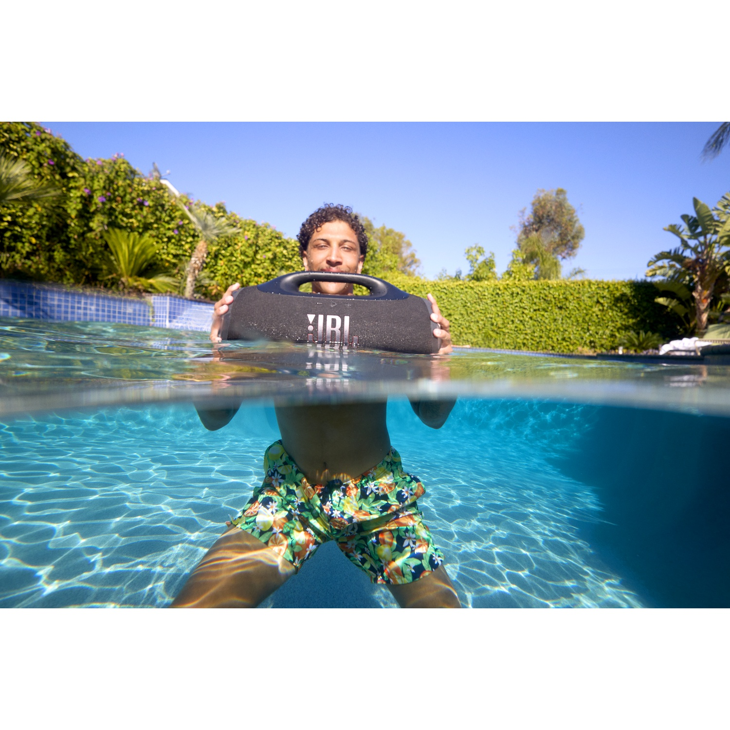 Man in pool holds a black JBL speaker. He wears floral shorts. Blue sky and greenery are in the background. Water is clear.