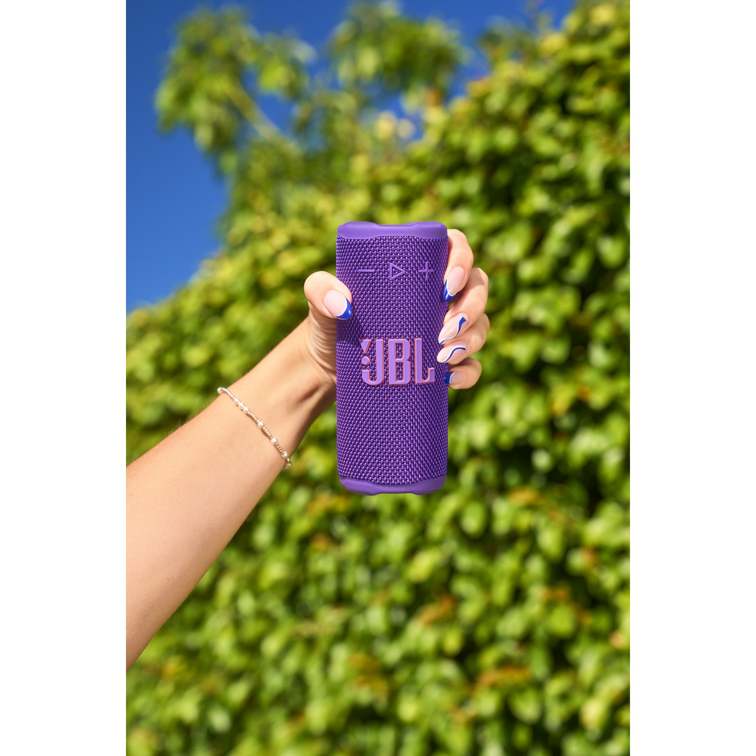 Une main tient un haut-parleur JBL violet sur un fond de feuillage vert et de ciel bleu. La main a des ongles à bout bleu.