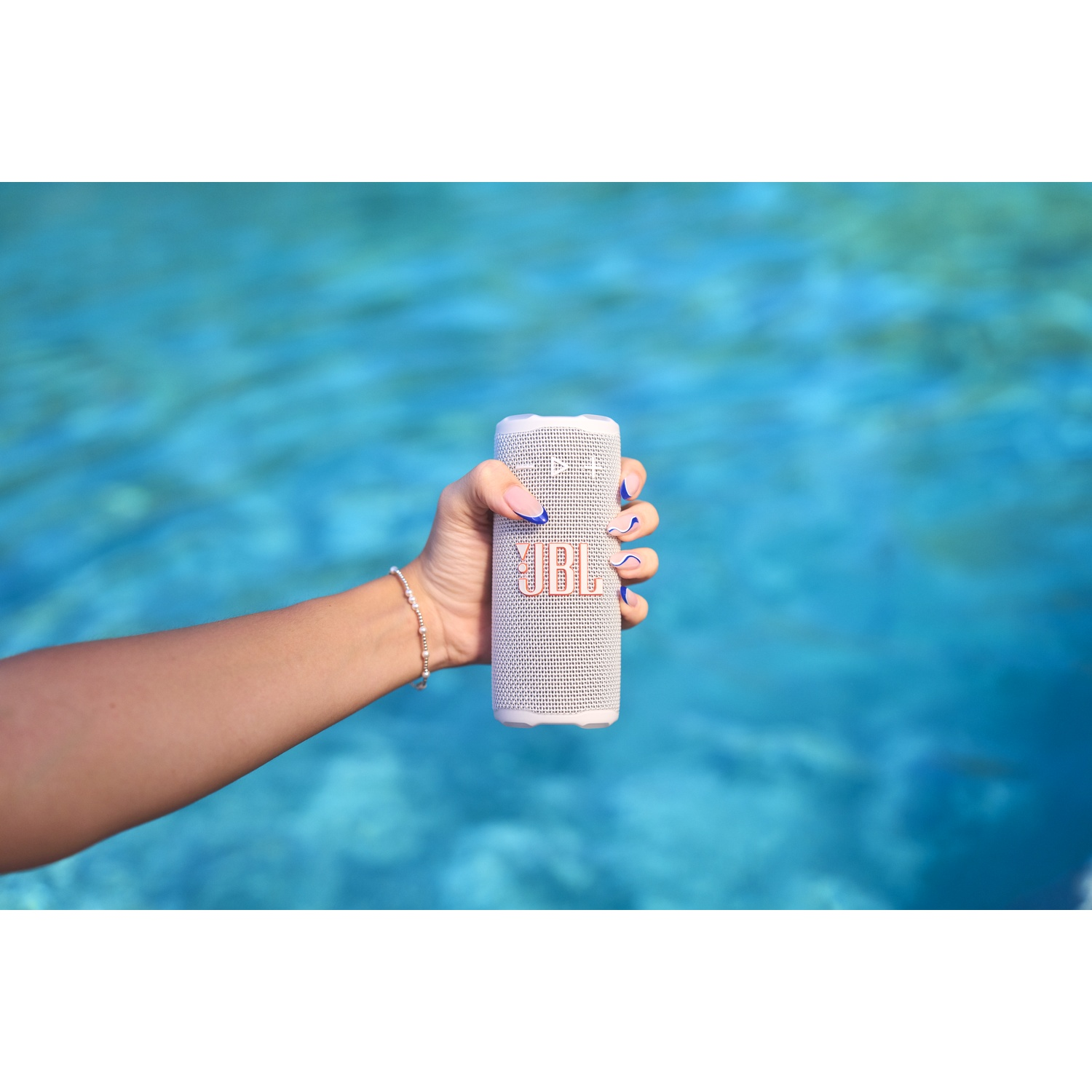Une main tient un haut-parleur JBL blanc avec des lettres corail, sur un fond d'eau bleue flou. La main a du vernis à ongles bleu.