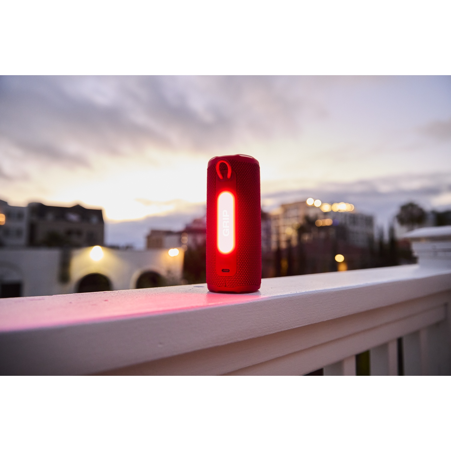 Un haut-parleur rouge avec une lumière vive est posé sur une balustrade blanche. Bâtiments et coucher de soleil en arrière-plan.