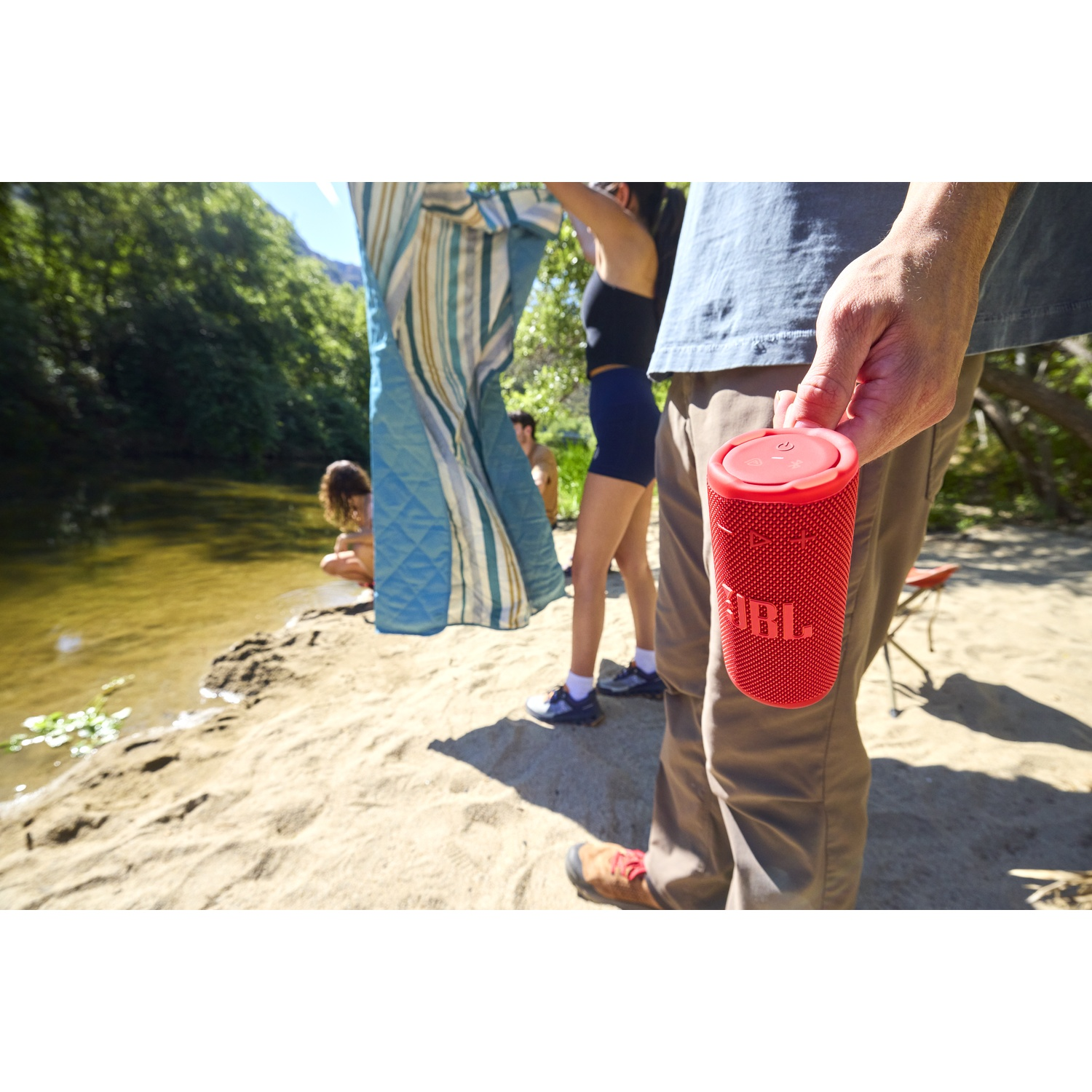 Une personne tient un haut-parleur JBL rouge sur une plage. Des personnes sont à l'arrière-plan, l'une tenant une couverture près d'une rivière.