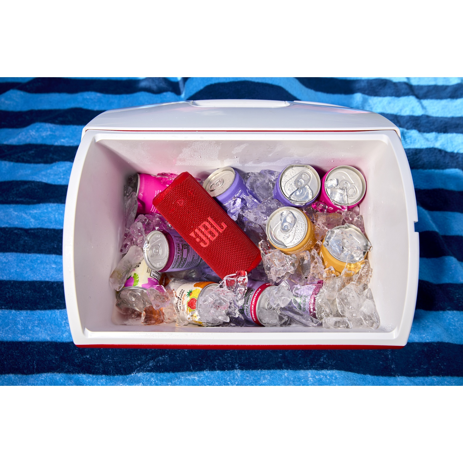 Une glacière remplie de glace et de canettes de soda. Un haut-parleur JBL rouge est à l'intérieur. La glacière repose sur une serviette rayée bleu et blanc.